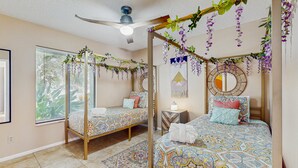 Una camera da letto con due letti, un ventilatore a soffitto, una finestra con persiane e piante decorative appese.