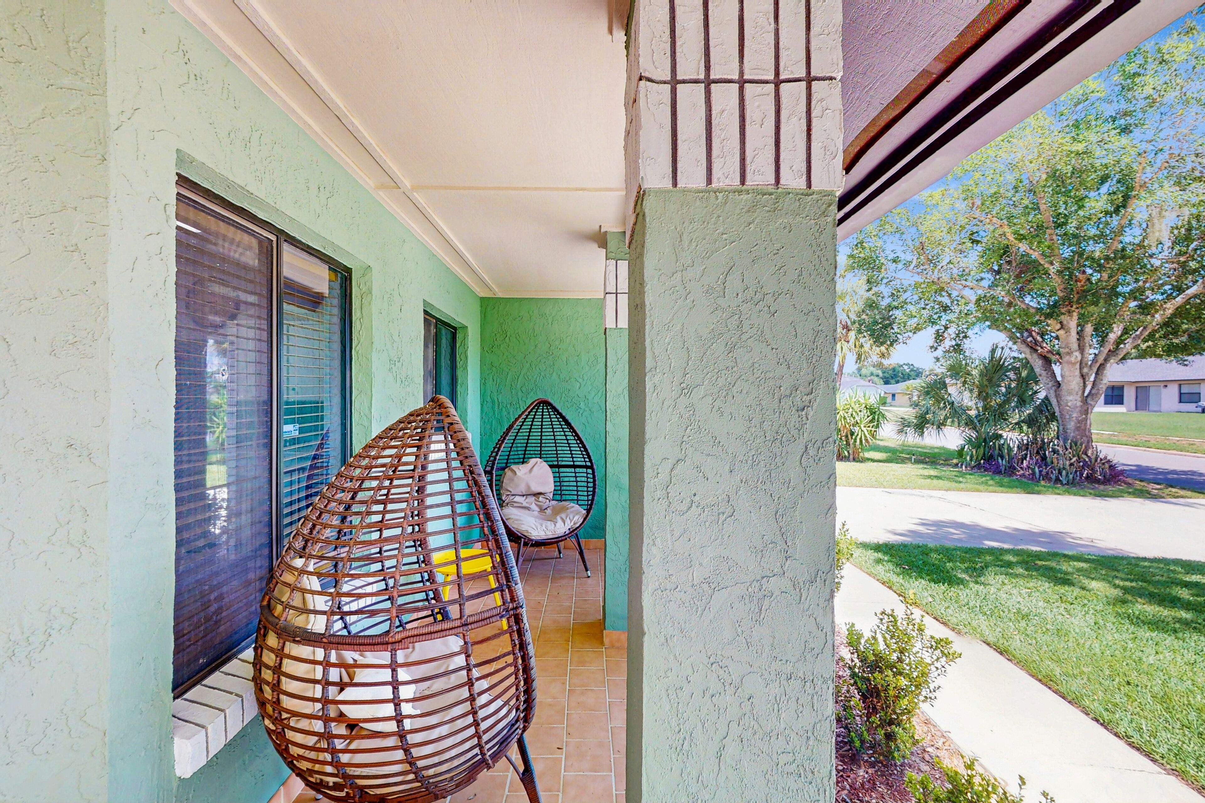 A covered porch with two wicker chairs, a small table, and a view of a tree-lined street.