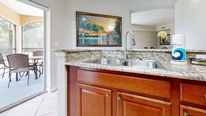 A kitchen with granite countertops, wooden cabinets, a stainless steel sink, and a painting above the sink. - Comfy Condo With Central AC, Wifi, Private Washer/dryer Minutes to Theme Parks (Orlando)