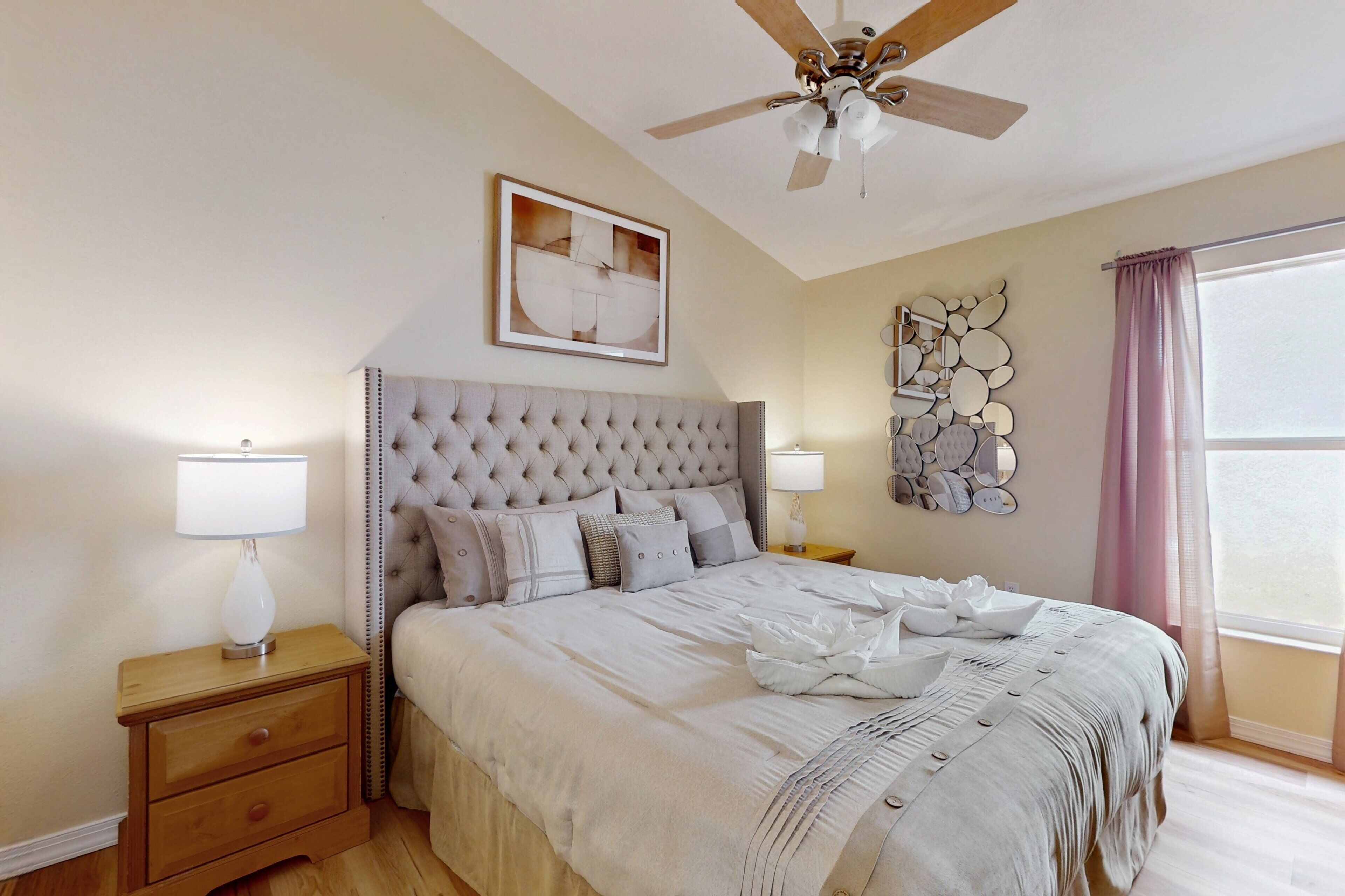 A bedroom with a tufted headboard, a ceiling fan, a bedside table with a lamp, a framed artwork above the bed, and a window with curtains.