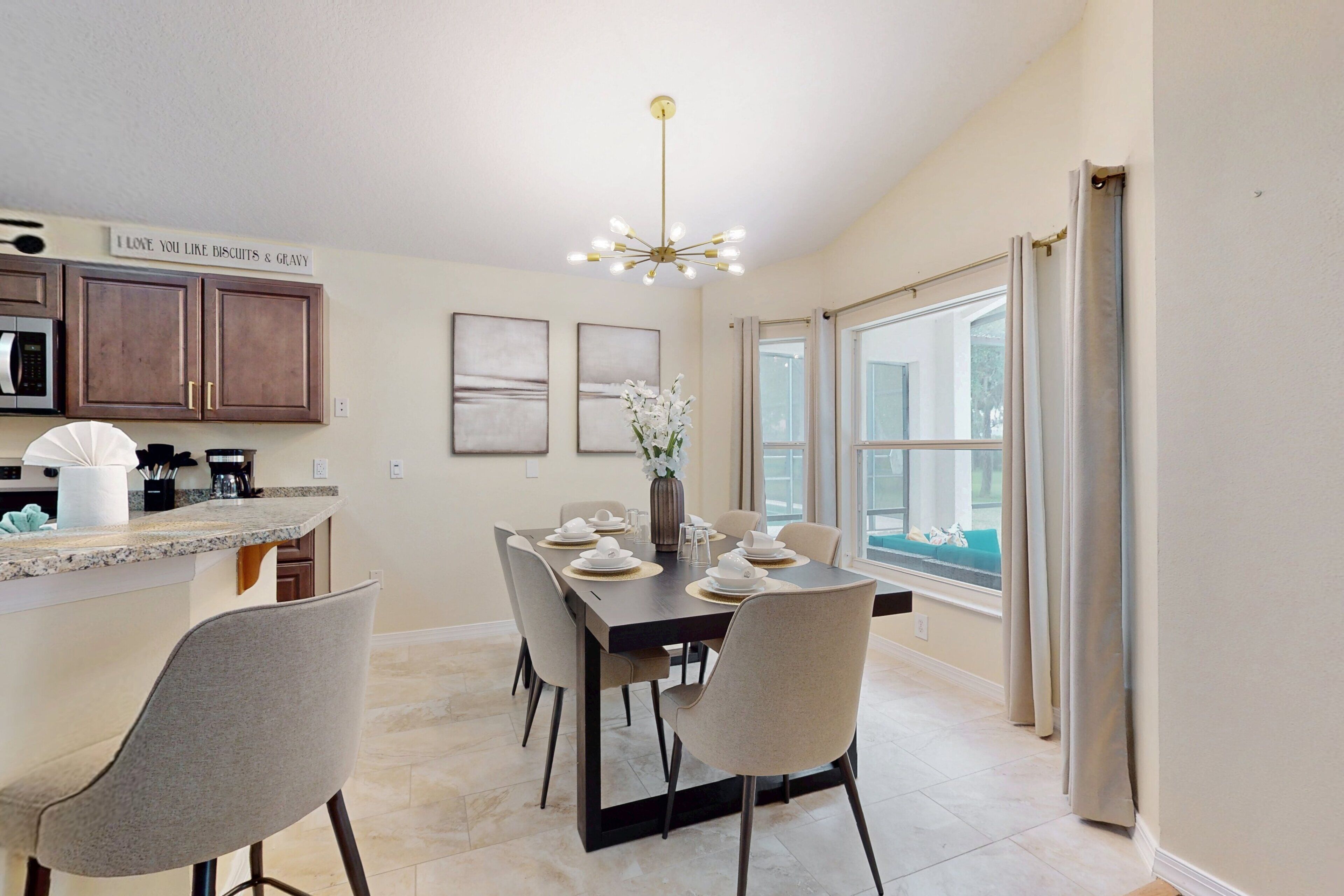 A dining area with a kitchen in the background, a chandelier, and a window with curtains.
