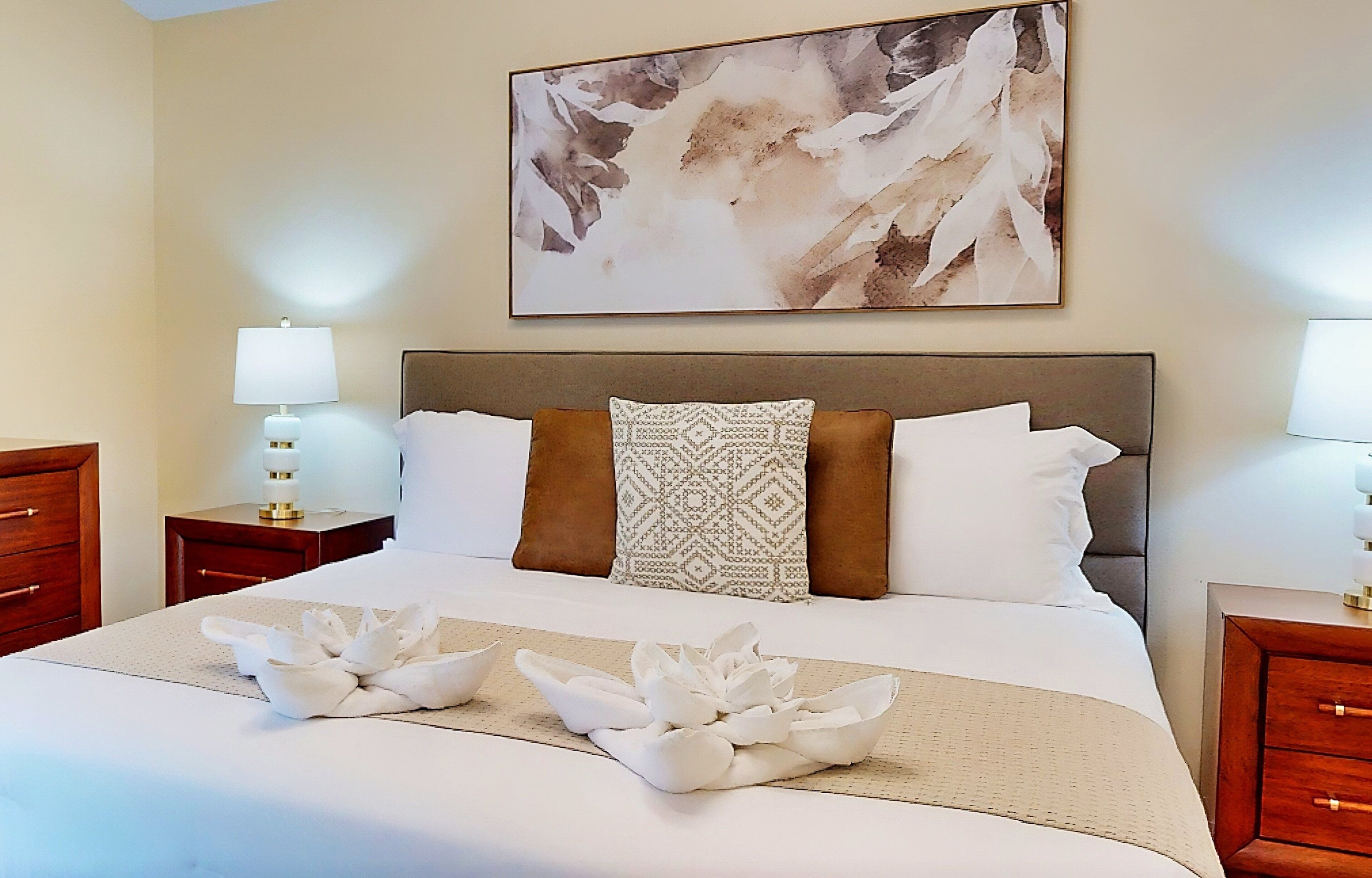 Una habitación de hotel con una cama grande, dos mesitas de noche con lámparas, un cojín decorativo y un cuadro en la pared.