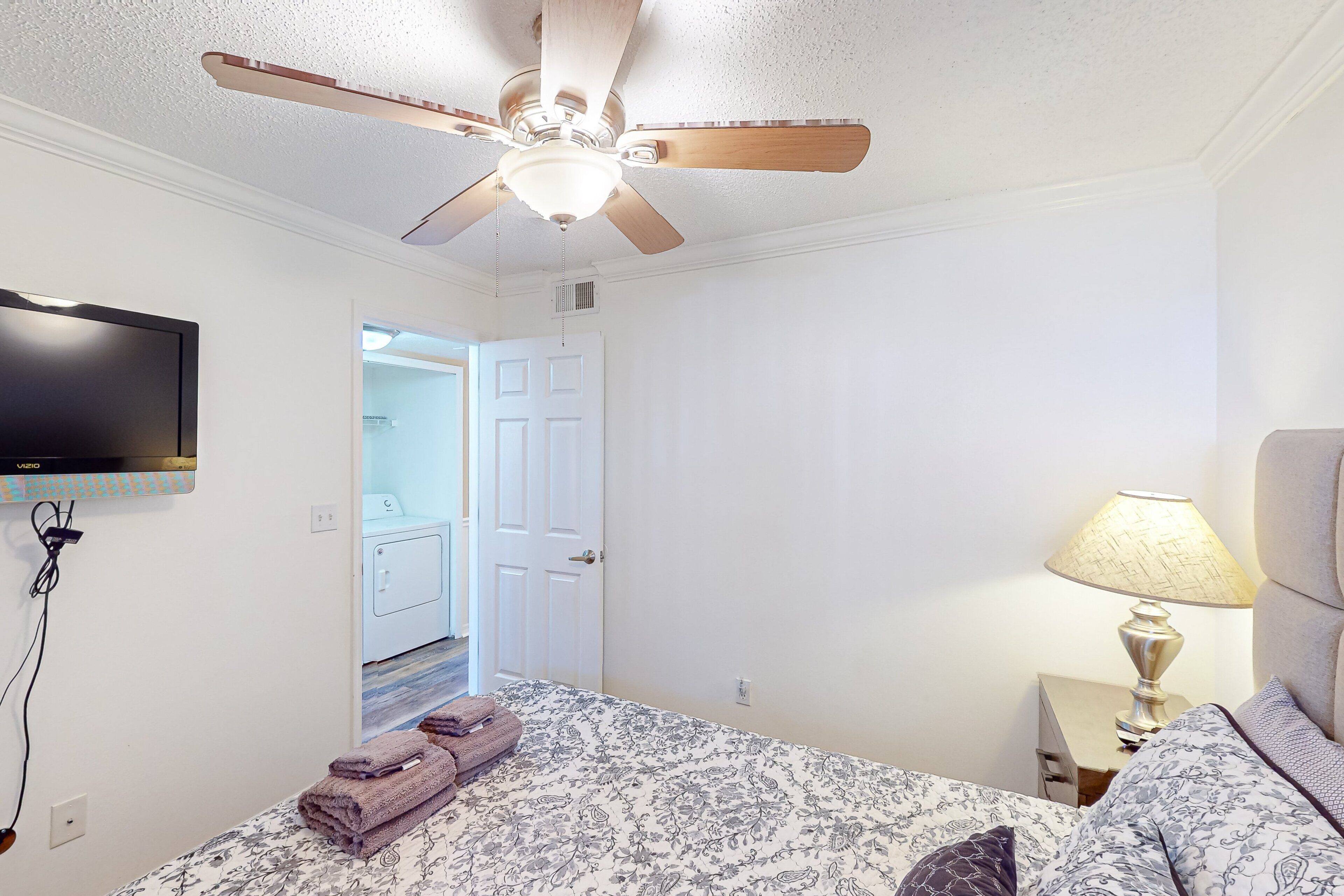 A hotel room with a bed, a ceiling fan, a TV mounted on the wall, a bedside lamp, and a door leading to a bathroom.