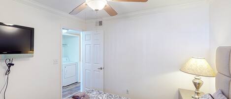 A hotel room with a bed, a ceiling fan, a TV mounted on the wall, a bedside lamp, and a door leading to a bathroom.