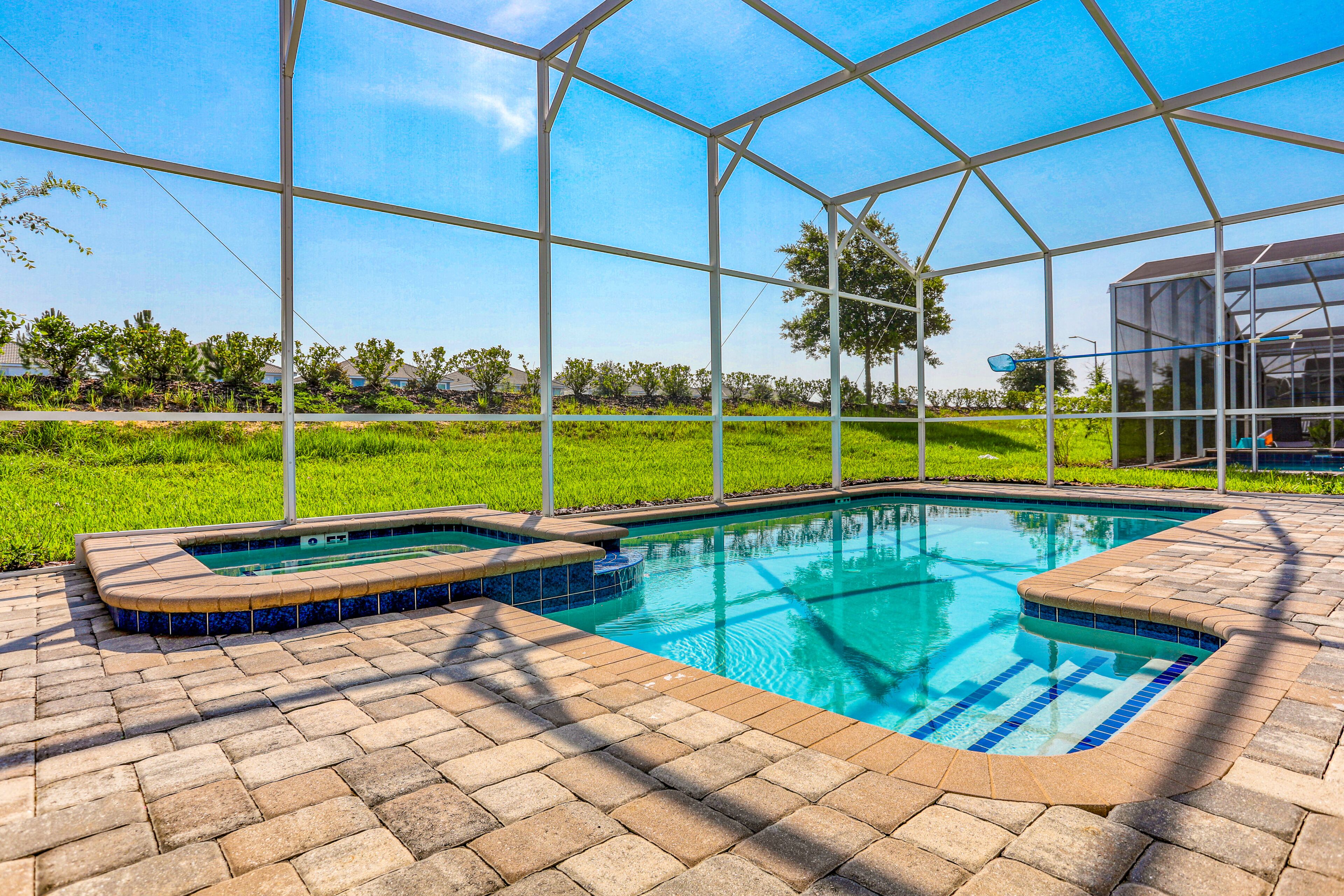 Ein Schwimmbad mit Glasüberdachung, umgeben von einer Ziegelterrasse, mit Blick auf ein grünes Feld und Bäume im Hintergrund.