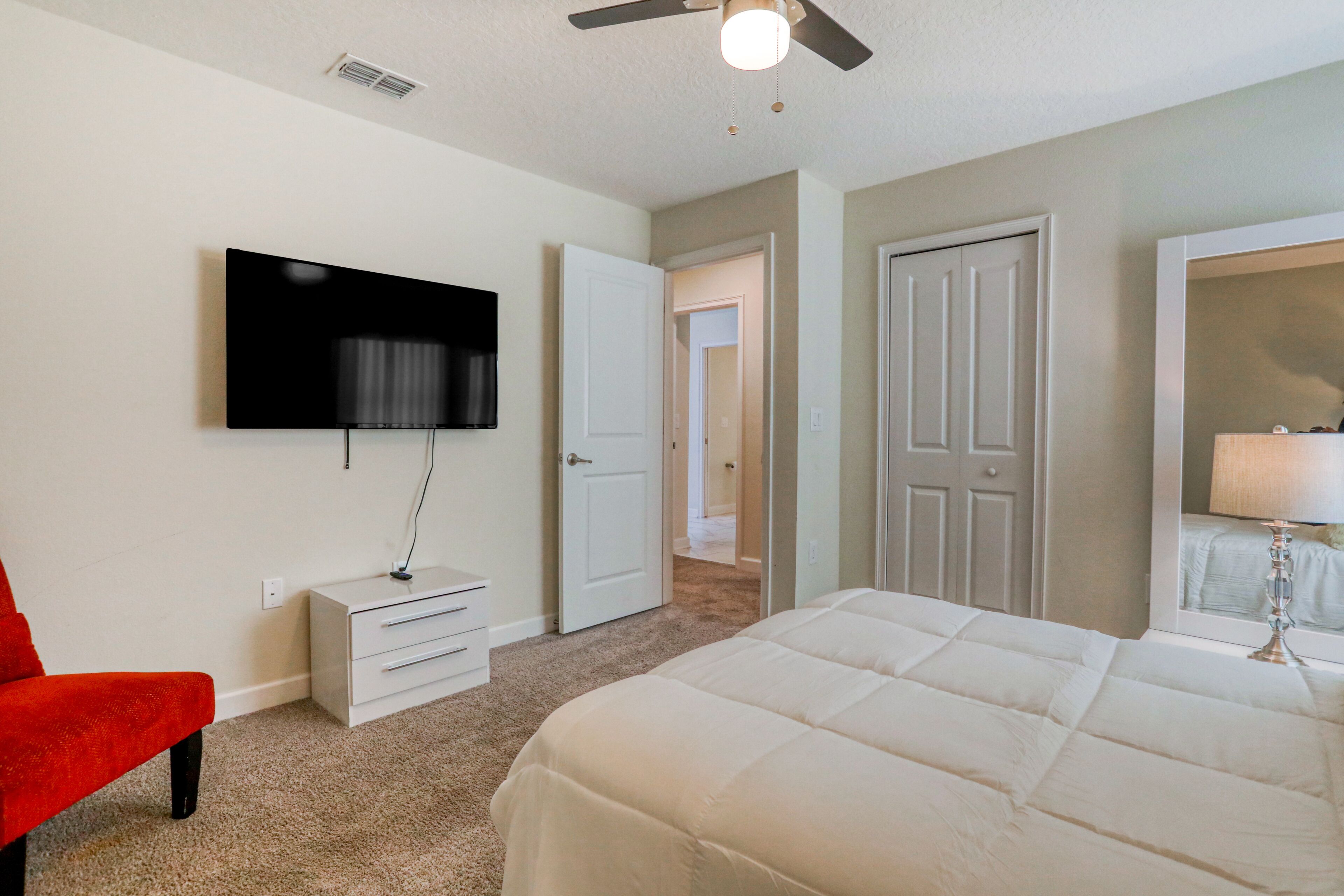 A bedroom with a bed, a TV mounted on the wall, a red chair, a white dresser, and a door leading to another room.