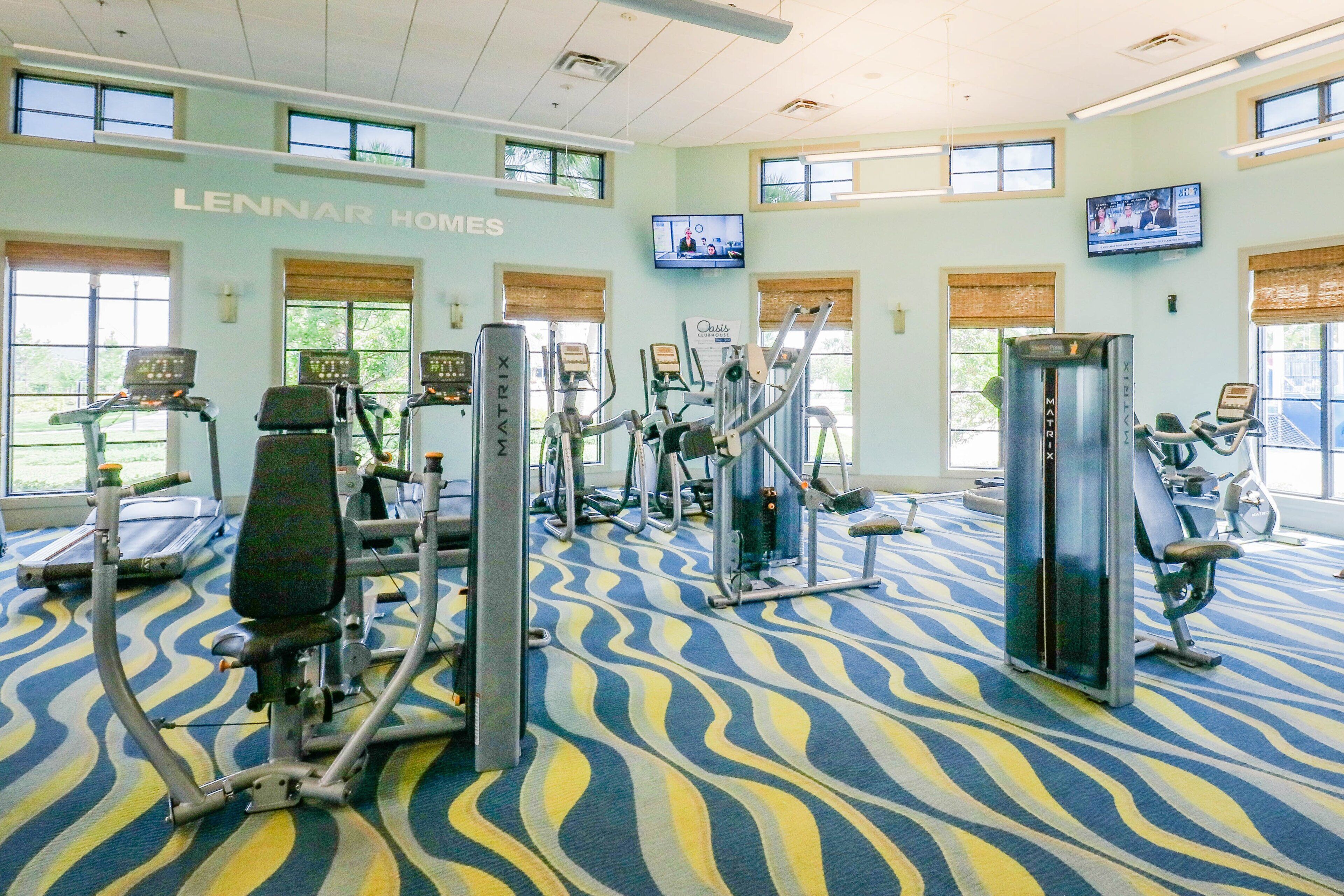 A gym with various exercise machines, large windows, and a patterned carpet.