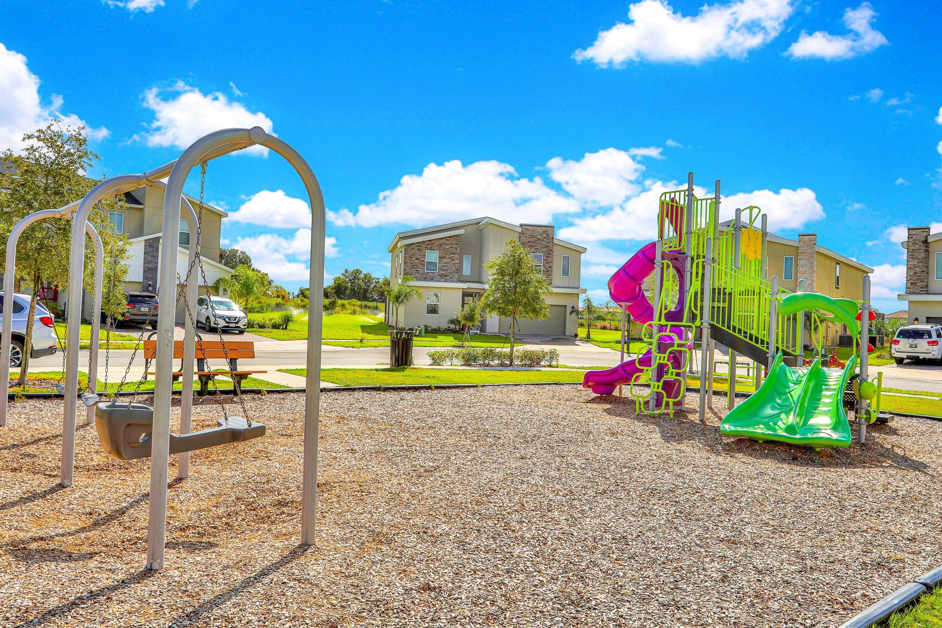 A playground with swings and slides, surrounded by a residential area with houses and parked cars.