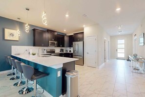 A modern kitchen with dark cabinets, a large island with bar stools, stainless steel appliances, and pendant lighting. - Luxury Resort Home With Themed Rooms, air Hockey, Pool Table, Pool and hot tub (Davenport)