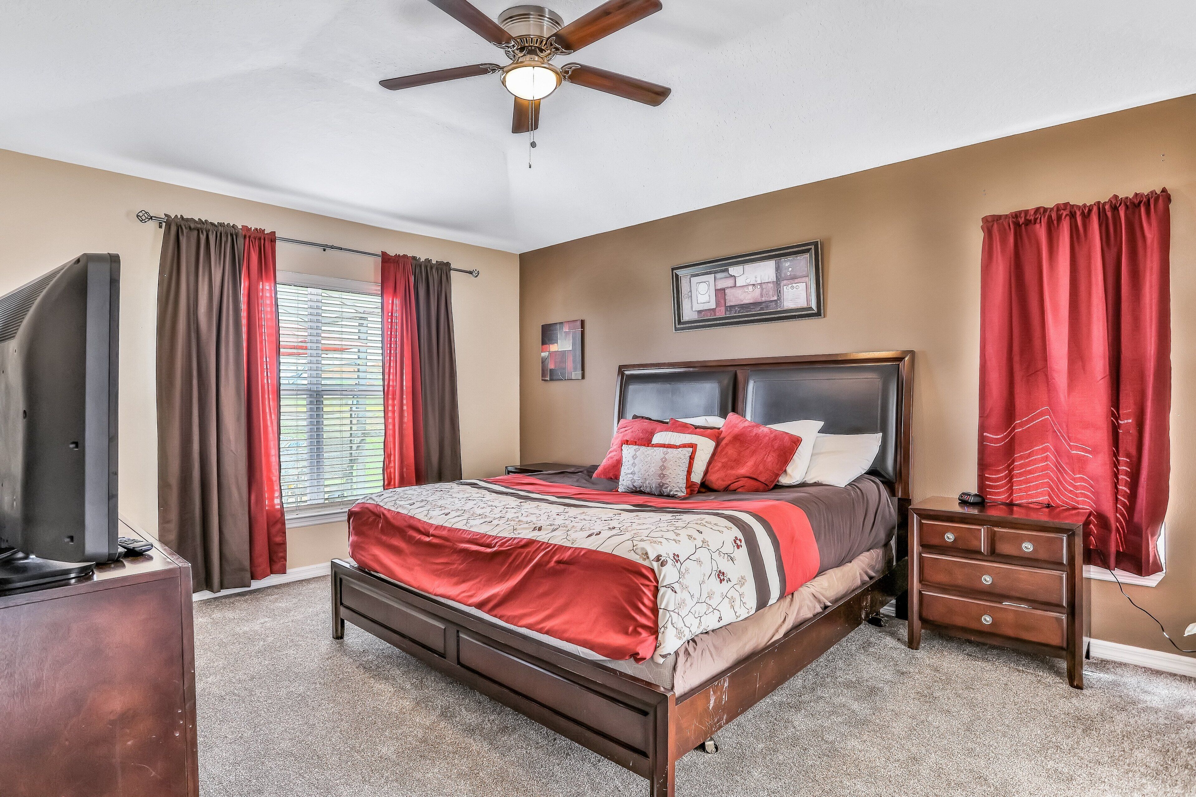 A bedroom with a bed, a nightstand, a ceiling fan, a TV, and a window with curtains.