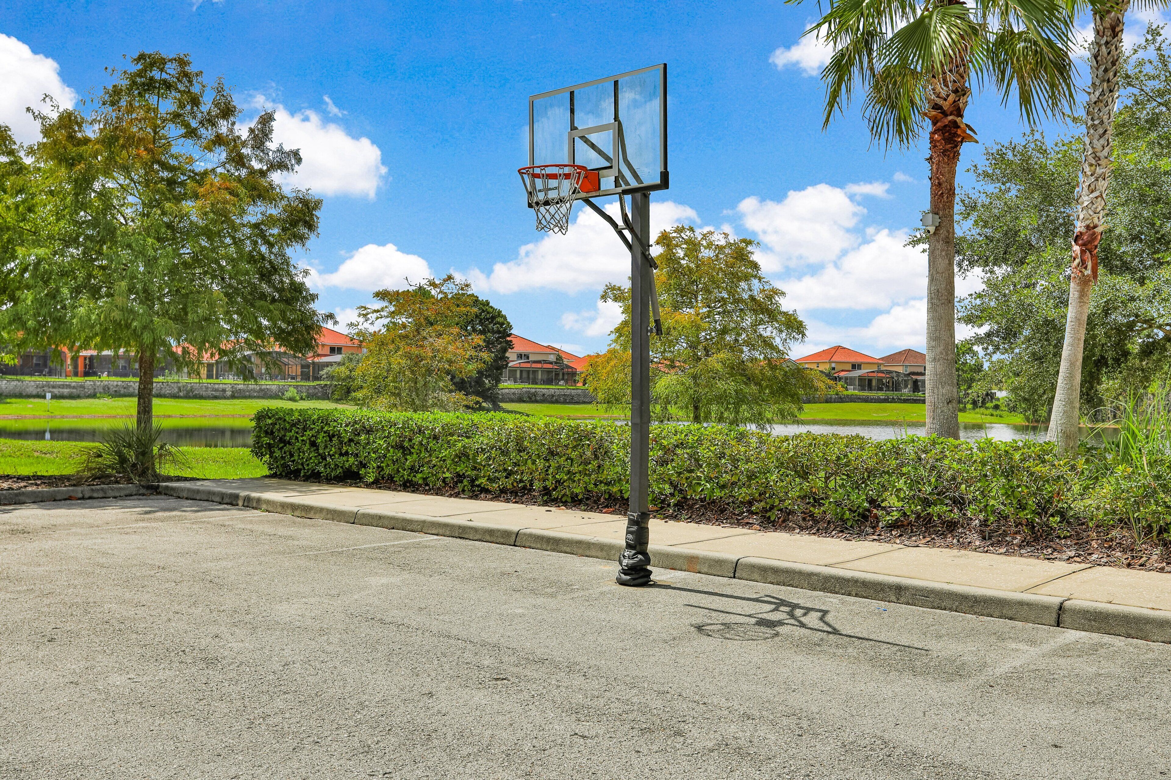Een basketbalring in een buitengebied met een verhard oppervlak, omgeven door groen en een heldere hemel.