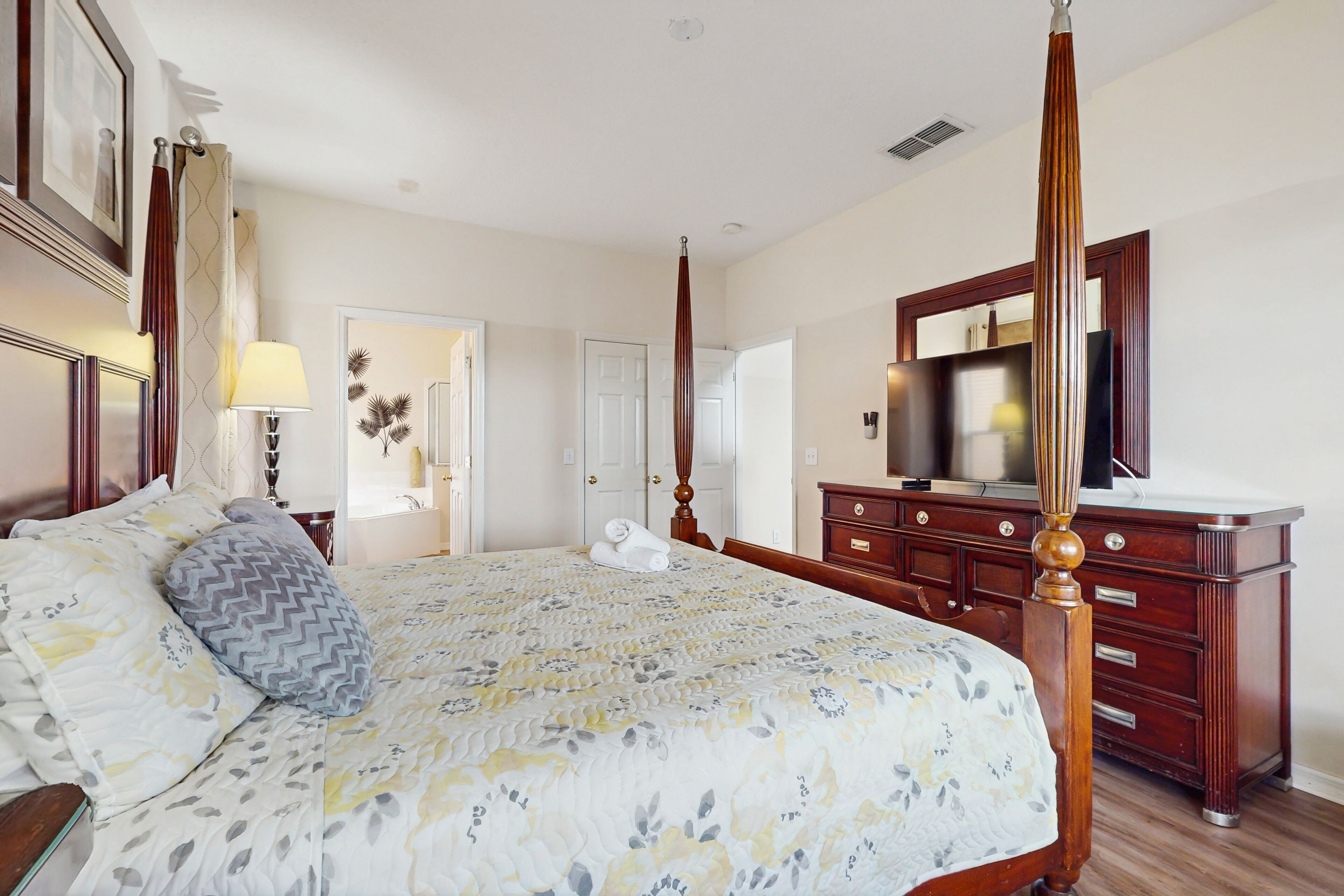 A bedroom with a four-poster bed, a wooden dresser, a television, and a door leading to another room.