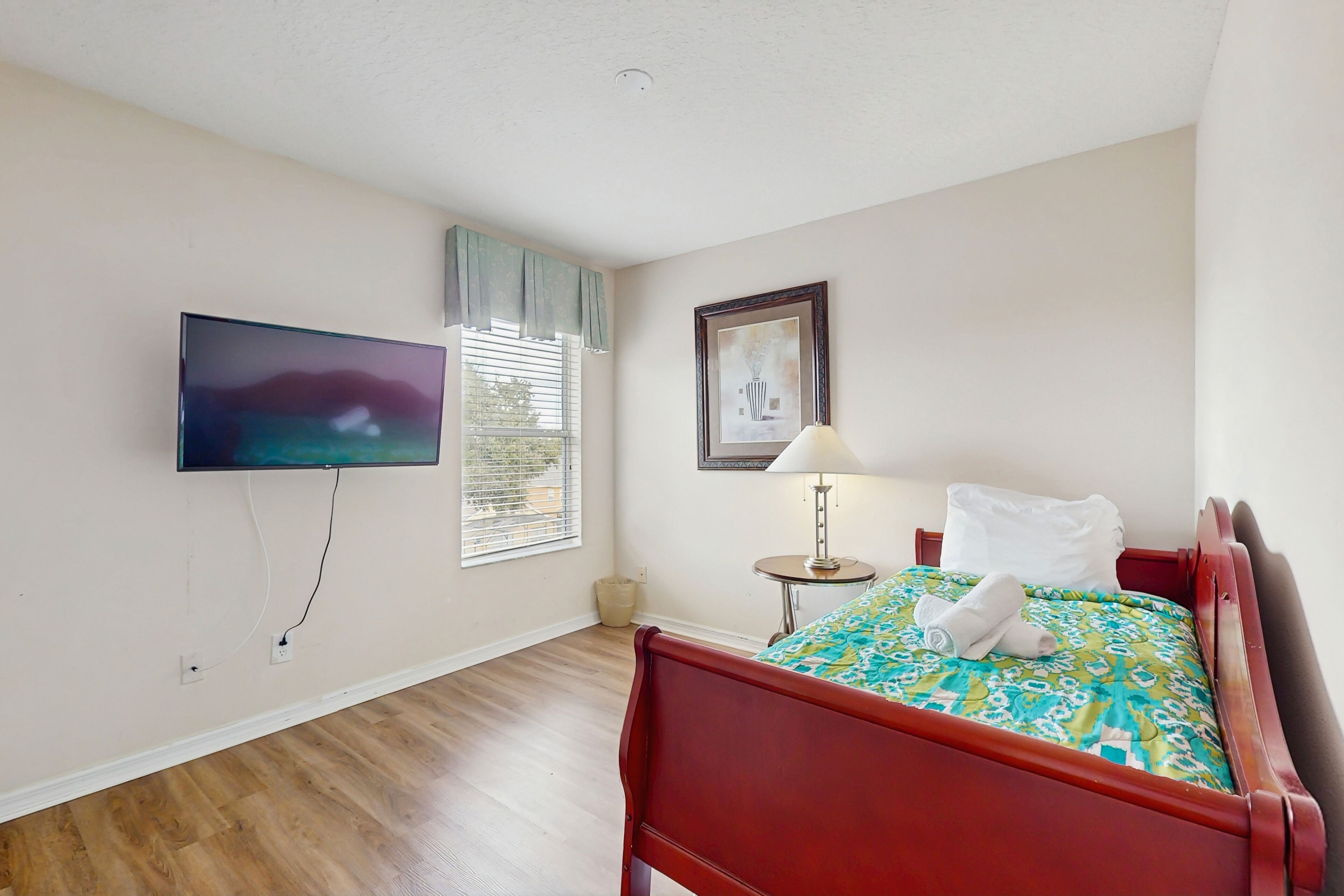 A bedroom with a wooden floor, a bed with a patterned bedspread, a television mounted on the wall, a window with blinds, and a framed picture on the wall.
