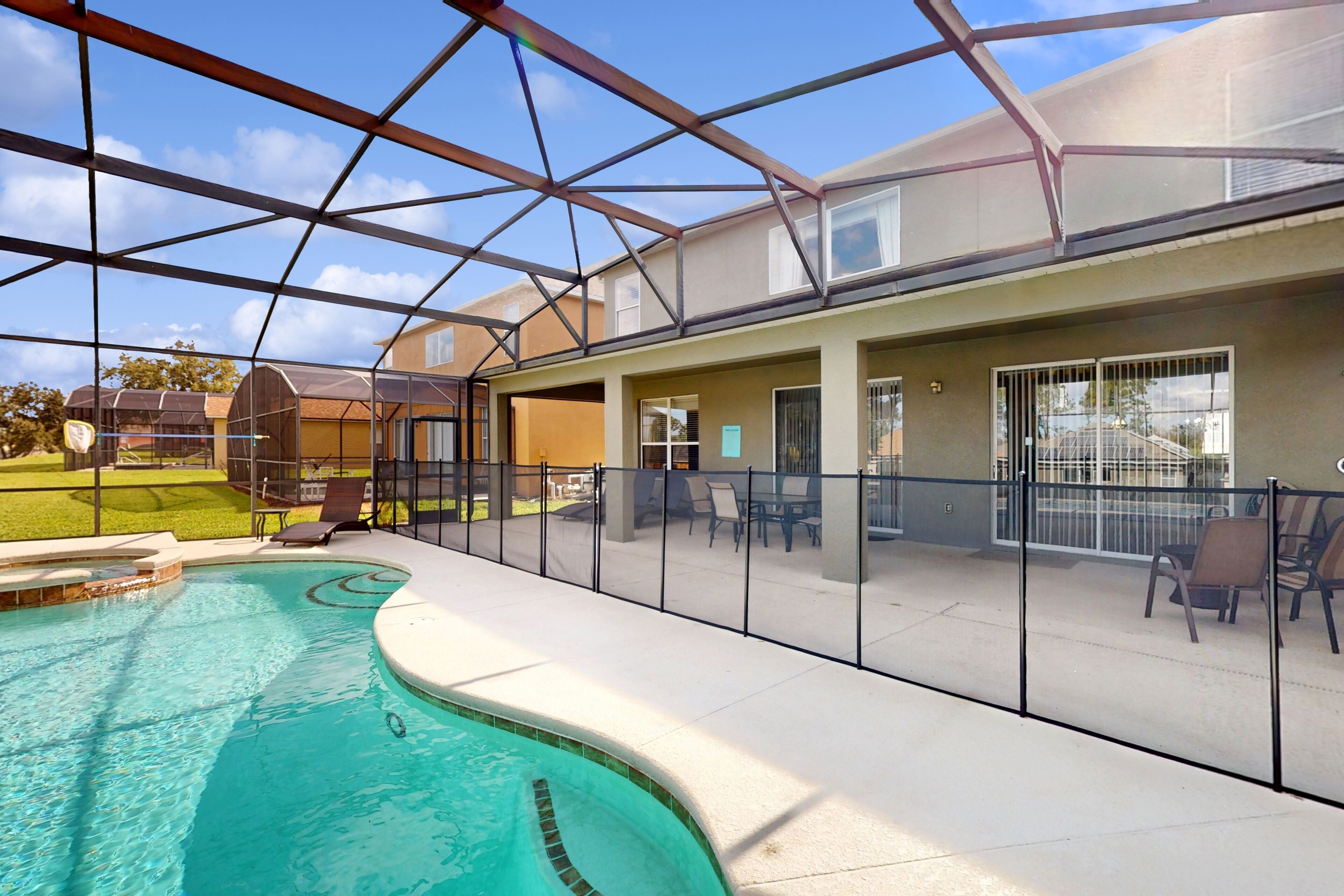 A covered outdoor swimming pool with a glass enclosure, adjacent patio area with outdoor furniture, and a view of the surrounding neighborhood.