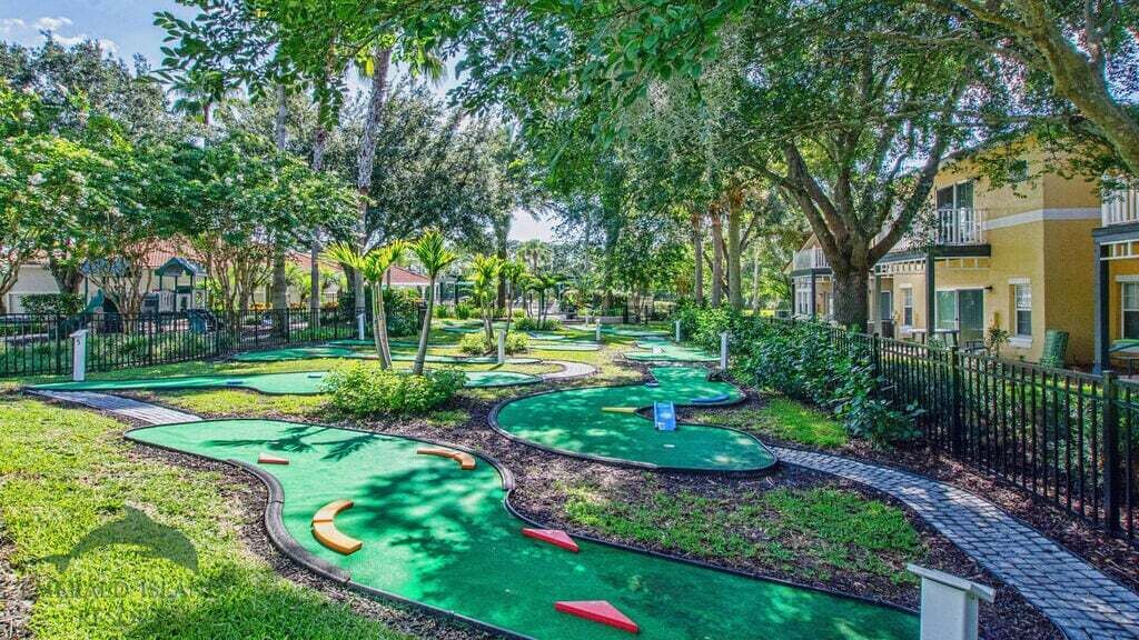 A miniature golf course with multiple green putting areas, surrounded by trees and a black fence.