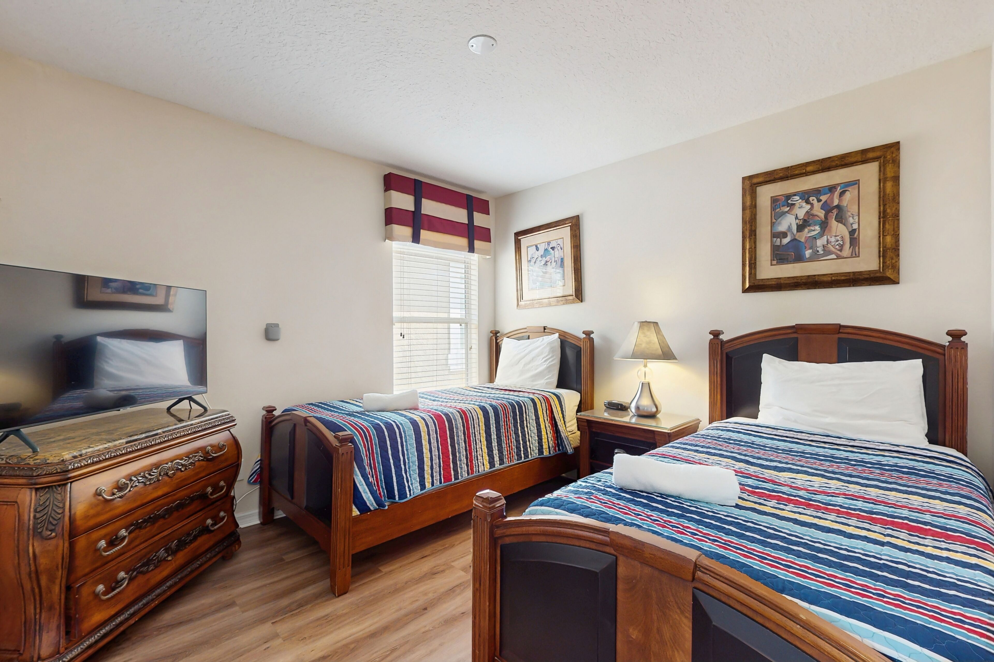 Une chambre d’hôtel avec deux lits en bois, une commode, un téléviseur et un tableau encadré au mur.