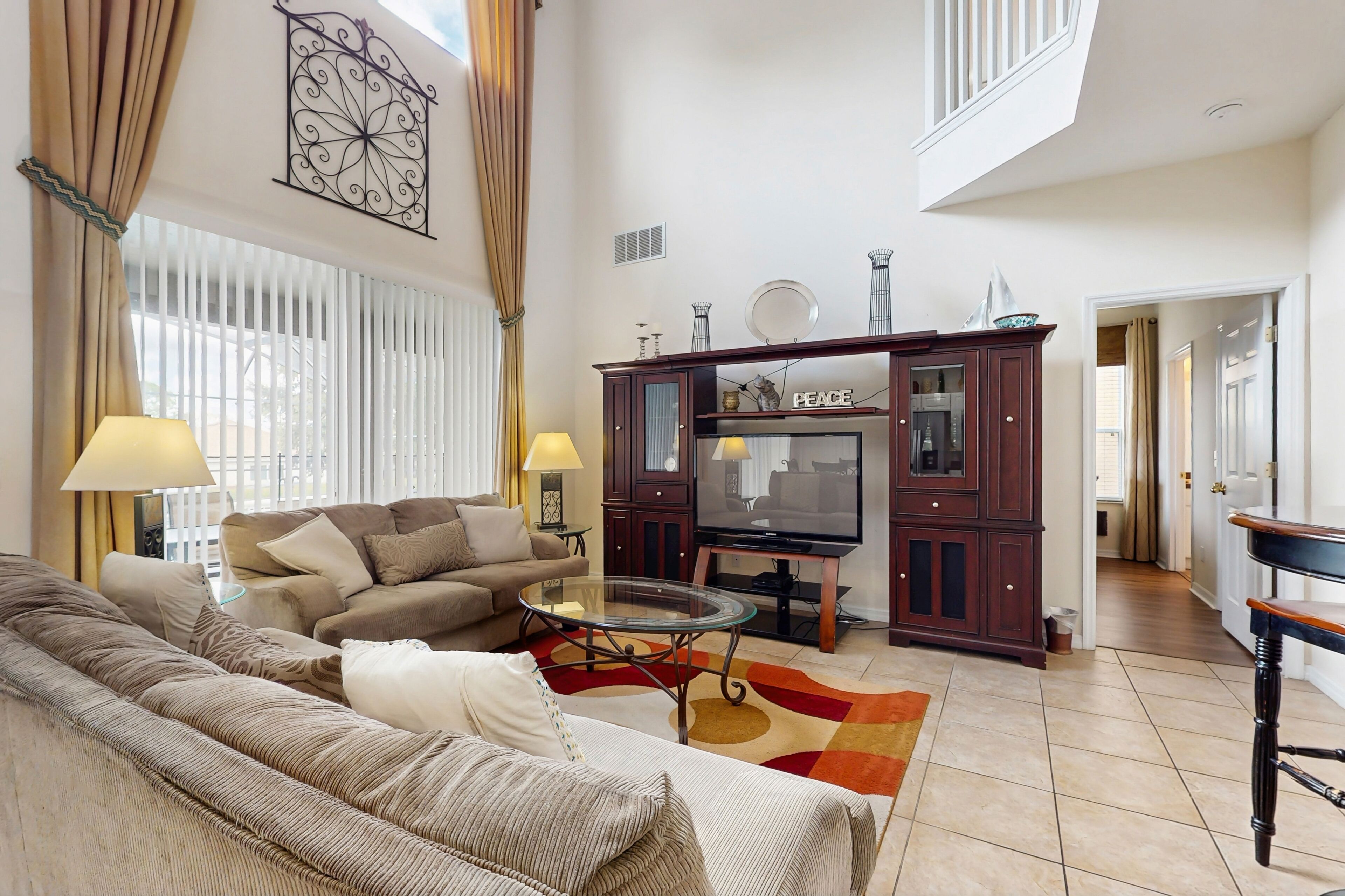 A spacious living room with a sofa set, a glass coffee table, and a TV. There is a wooden cabinet with decorative items and a large mirror.