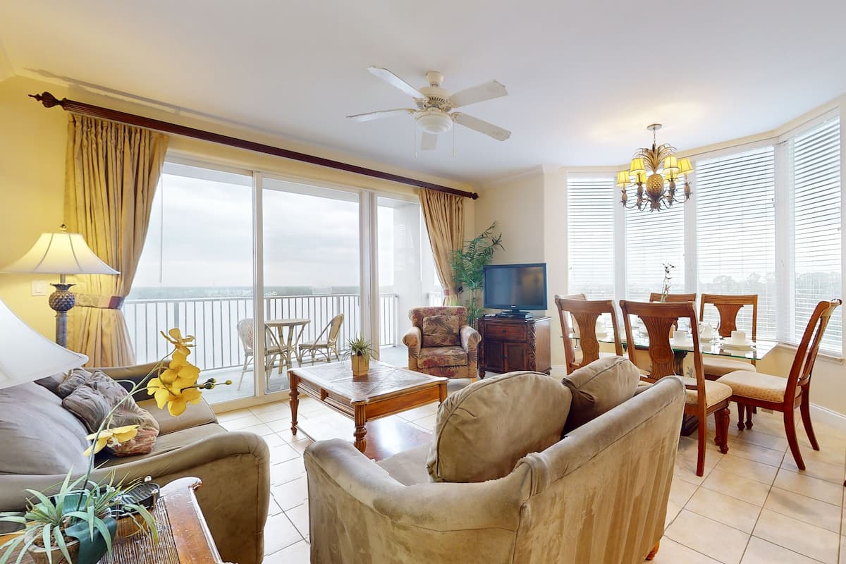 A spacious living room with a sofa, armchairs, a coffee table, and a television. There is a balcony with a view, a chandelier, and a dining area with chairs and a table.