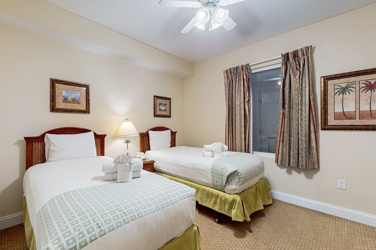 A hotel room with two beds, a ceiling fan, a window with curtains, and framed pictures on the walls.