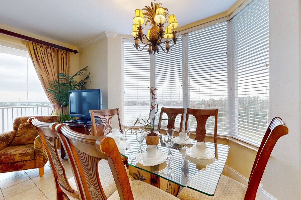 Un comedor con mesa de vidrio, sillas, candelabro, televisor y vistas amplias por las ventanas.
