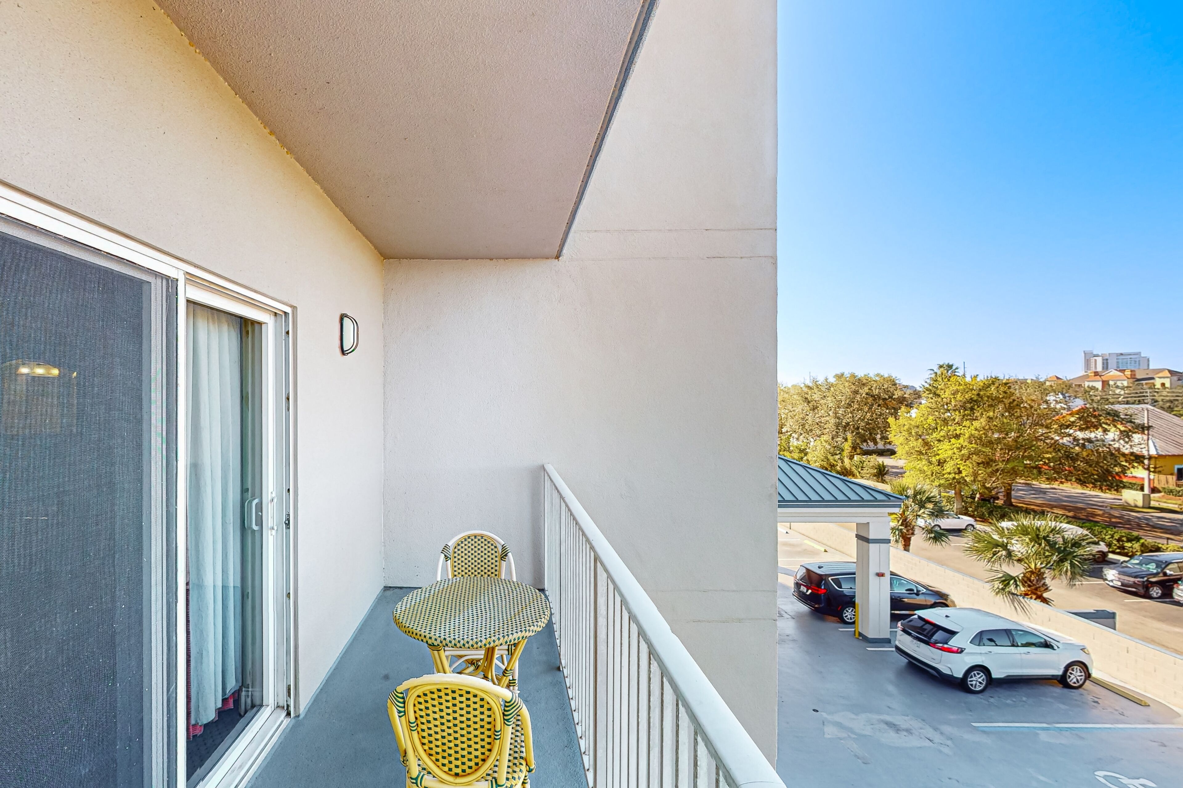 A balcony with two wicker chairs and a table, overlooking a parking area with cars and buildings.