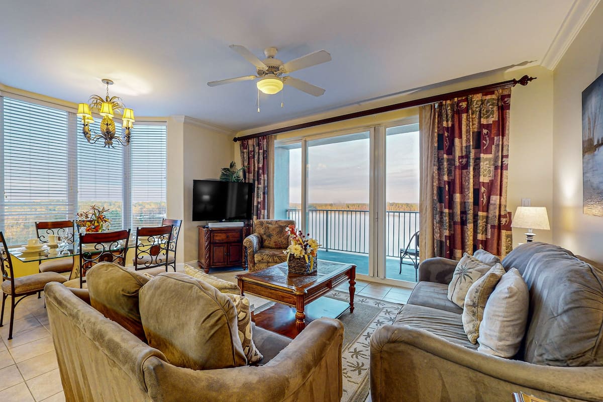 A spacious living room with a ceiling fan, a flat-screen TV, a dining area with a glass table, and a balcony with curtains.