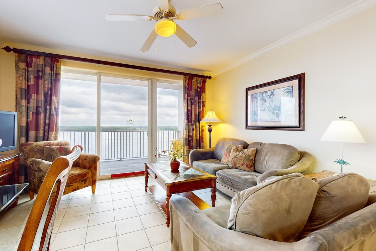 A living room with a sofa, armchair, and a television. There is a balcony with curtains and a ceiling fan.