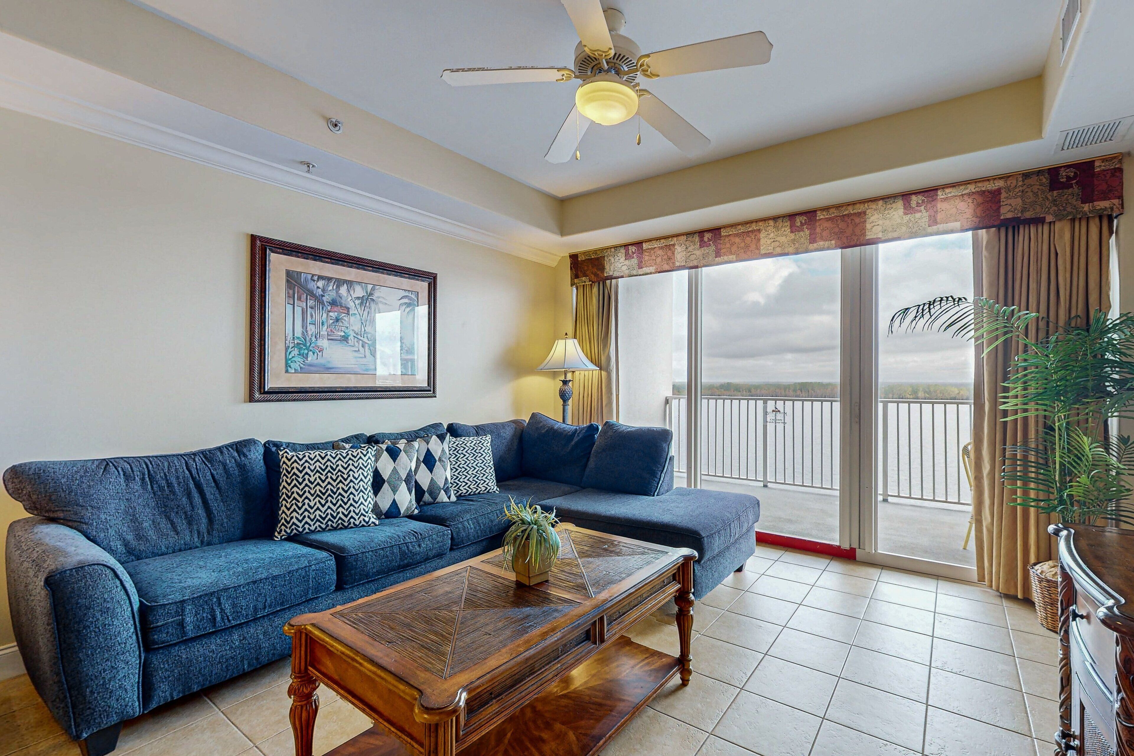 A living room with a blue sofa, a wooden coffee table, a ceiling fan, a large window with curtains, and a framed picture on the wall.