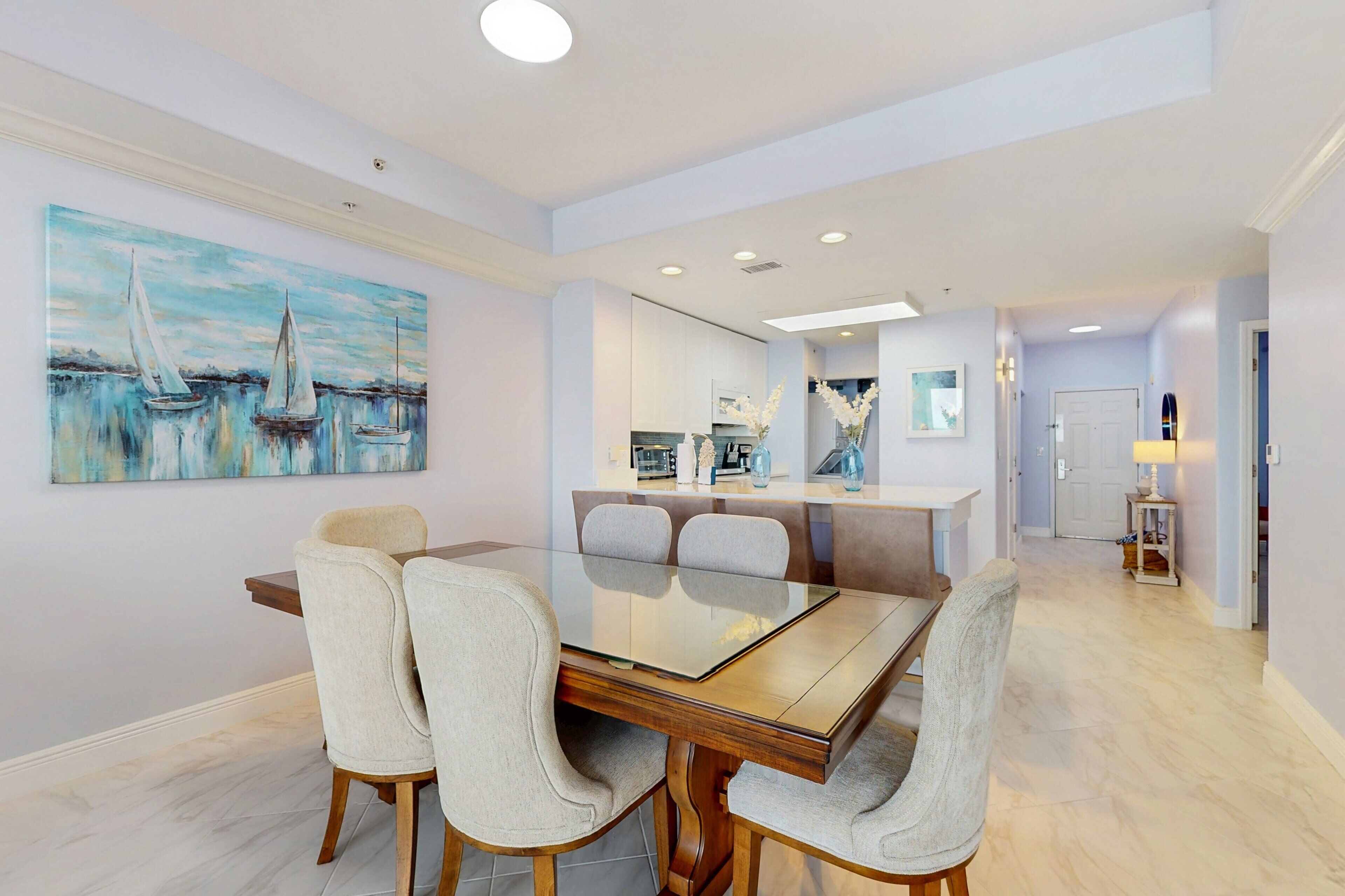 A modern dining area with a glass-top table, cushioned chairs, and a painting of sailboats on the wall.