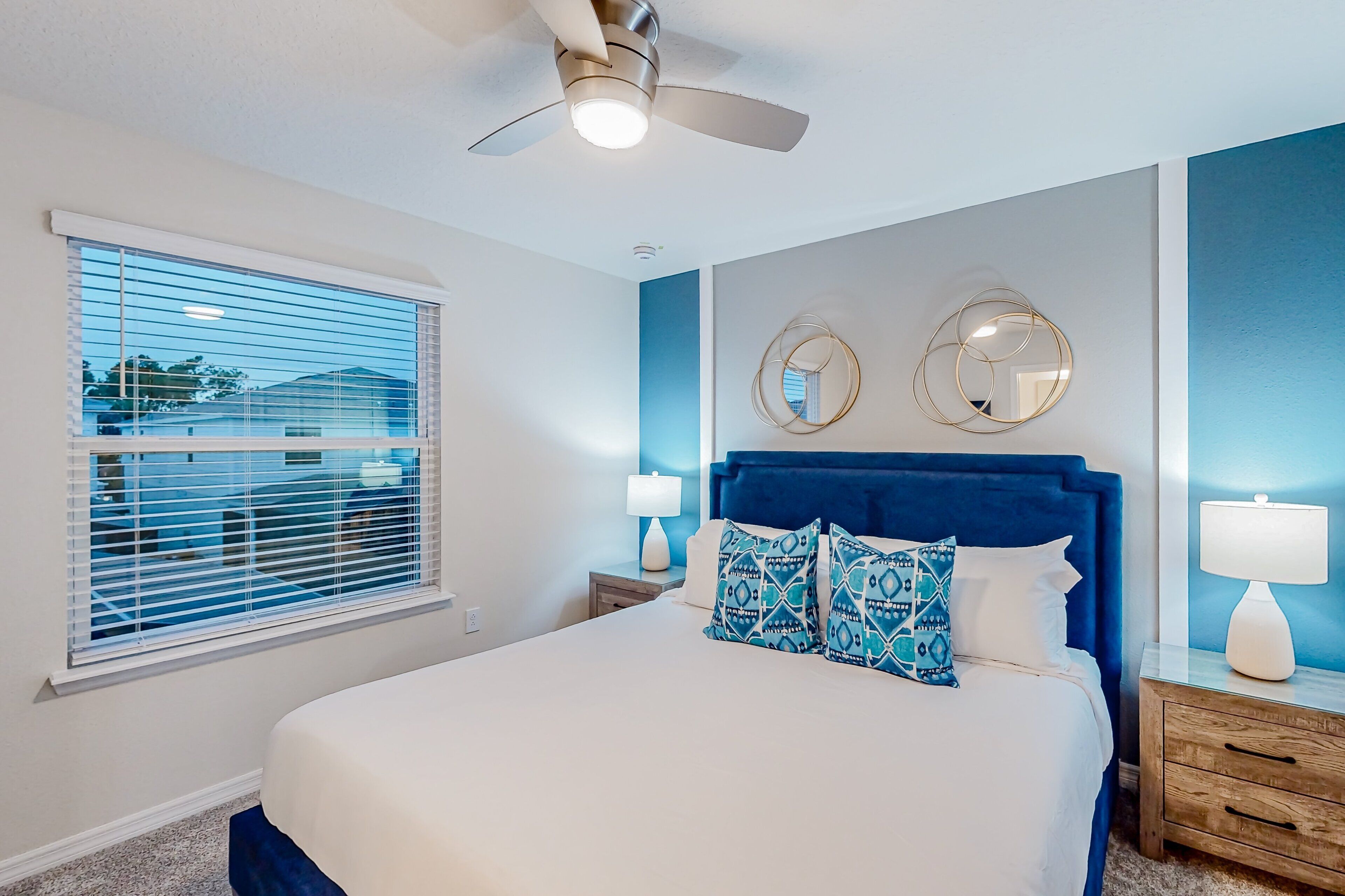 A bedroom with a bed, a ceiling fan, a window with blinds, a nightstand with a lamp, and two wall mirrors.