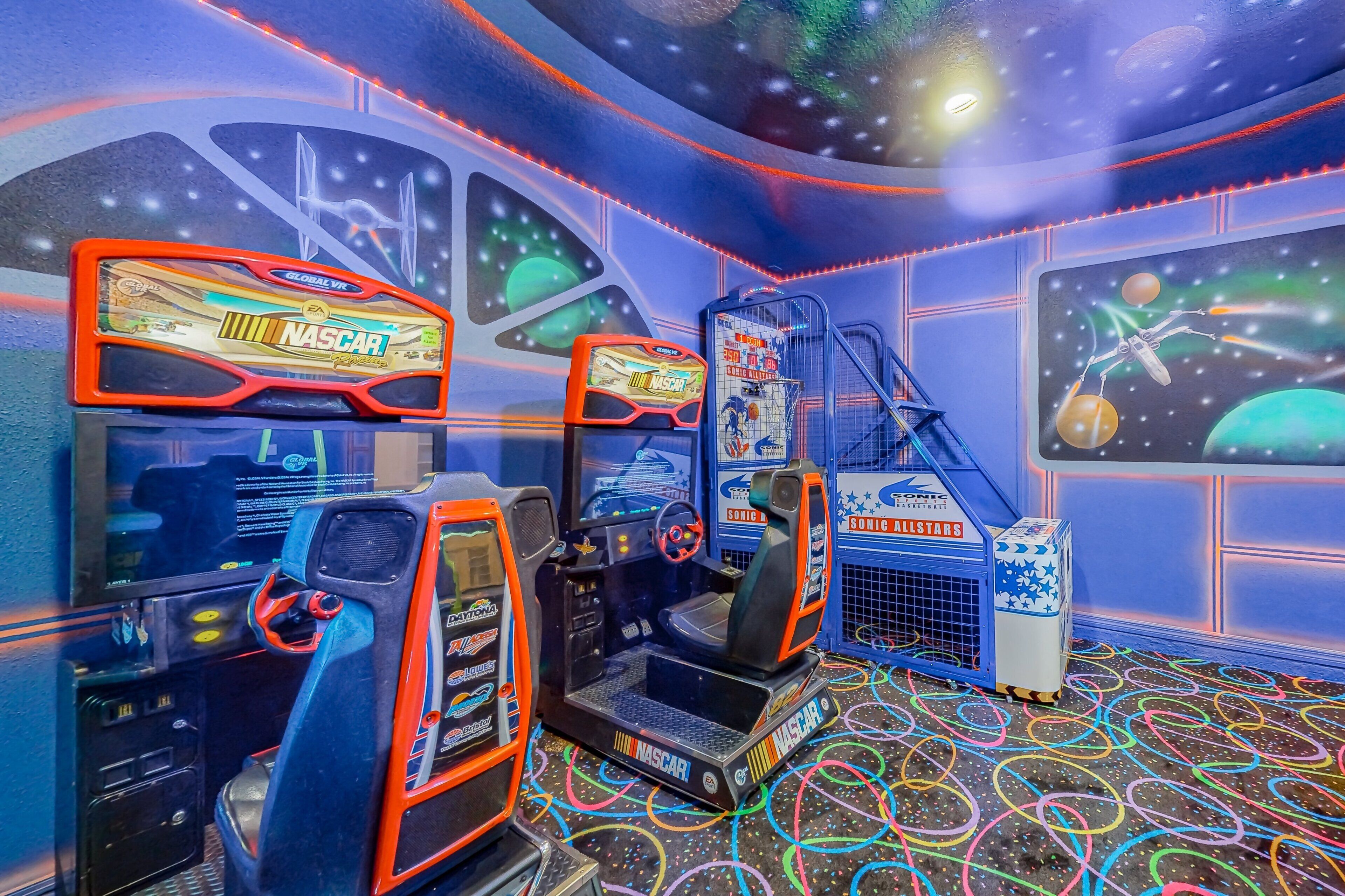 A virtual reality gaming area with multiple racing game stations, including NASCAR and Sonic Allstars, in a room with a starry ceiling and colorful floor design.