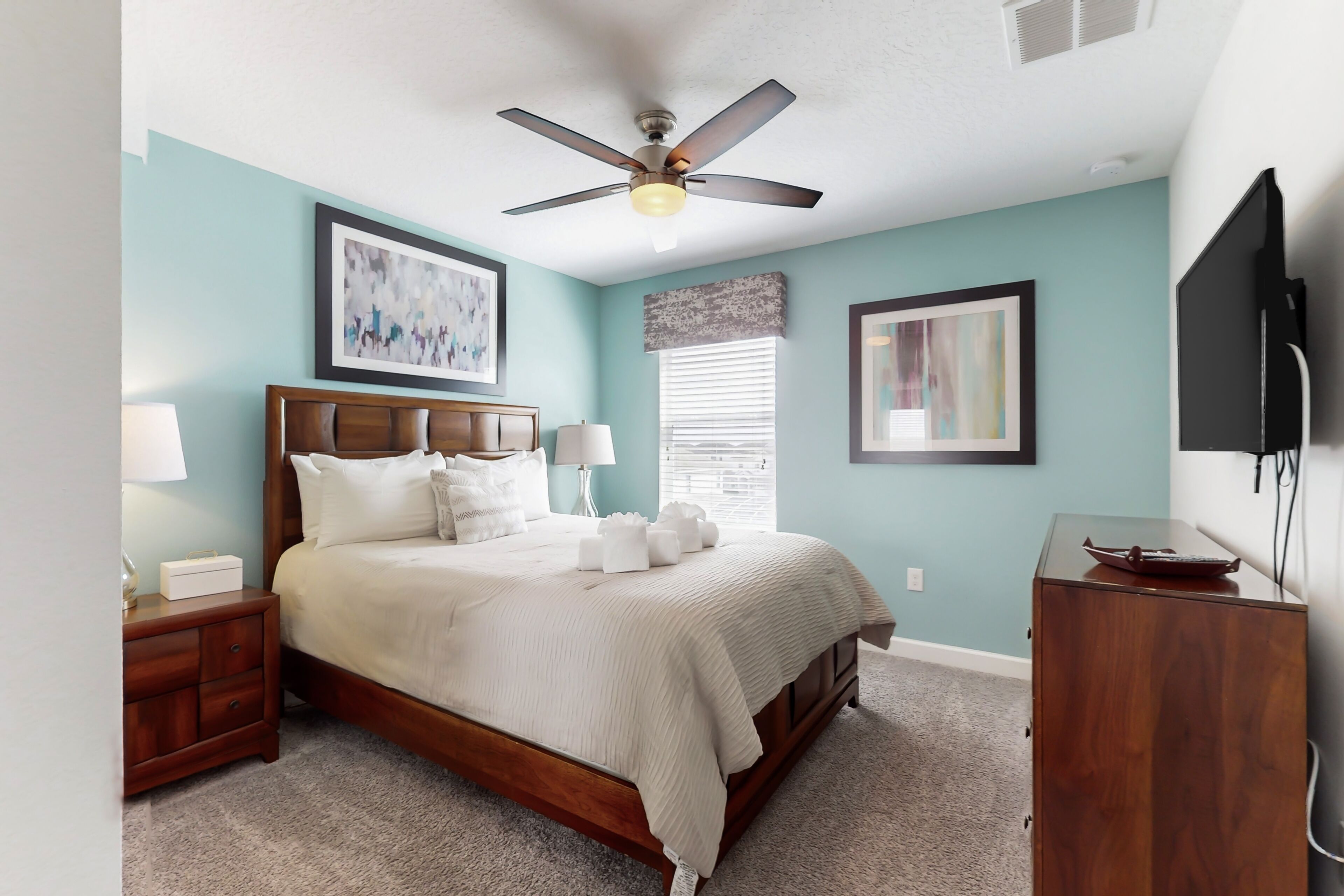 A bedroom with a bed, nightstands, a wooden dresser, a ceiling fan, and artwork on the walls.