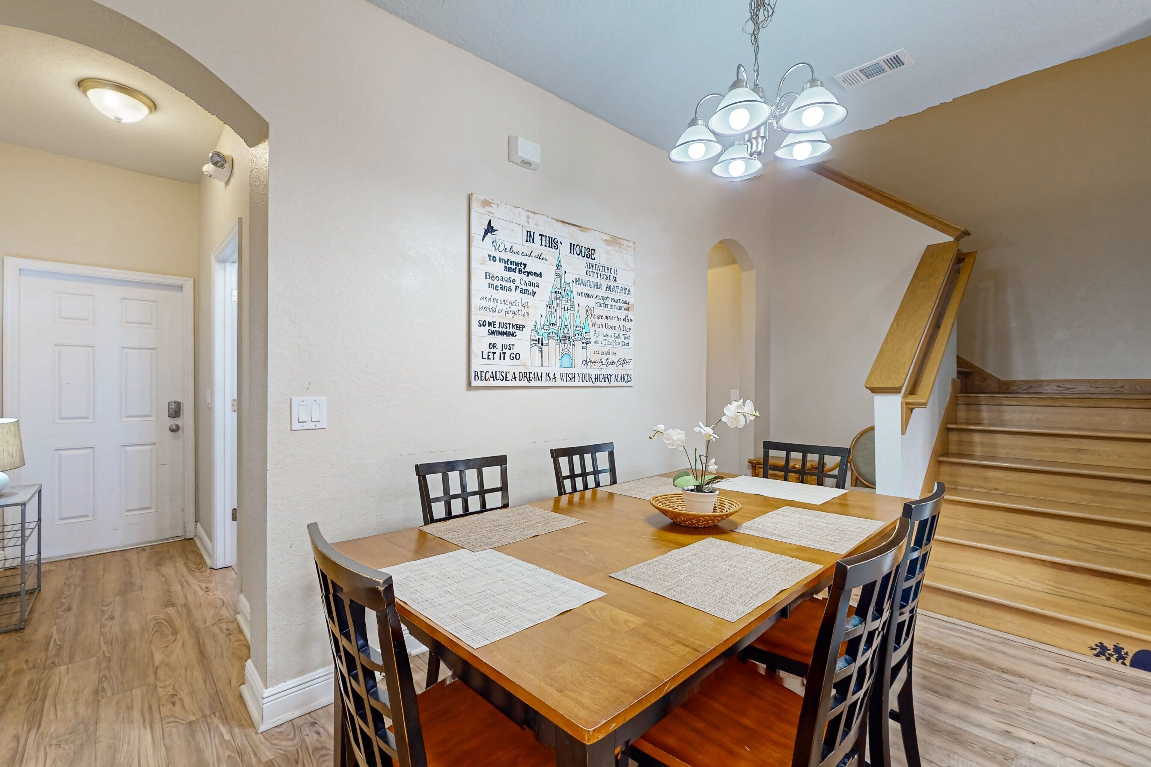 A dining area with a wooden table, four chairs, a chandelier, and a staircase.