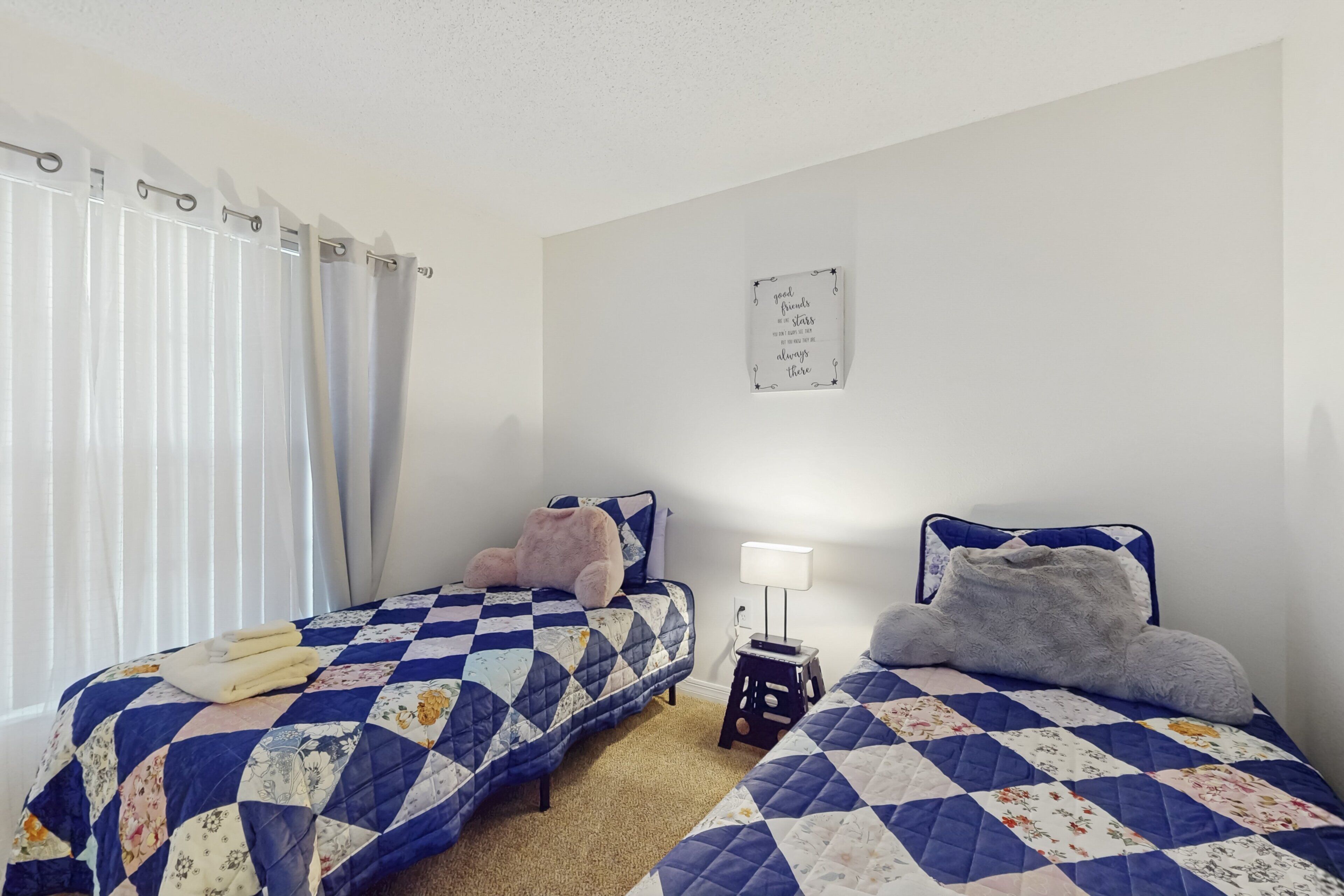 A bedroom with two beds, a nightstand, a wall-mounted sign, and a window with curtains.