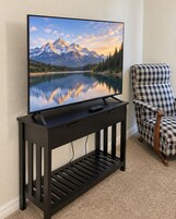 Un televisor de pantalla plana sobre un mueble negro, con un control remoto, ubicado en una habitación con una silla a cuadros y suelo alfombrado.