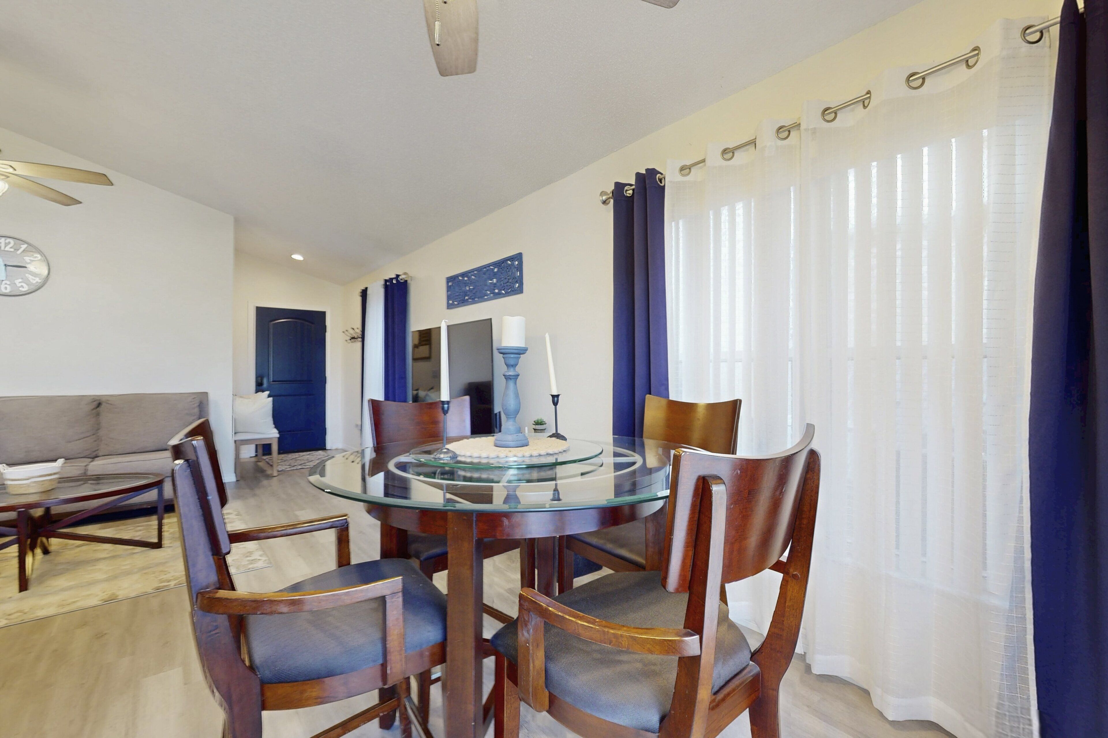 Un coin repas avec une table à plateau en verre, des chaises en bois, une horloge au mur et une porte visible au fond.