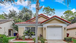 A residential house with a red-tiled roof, a garage, and a well-maintained front yard with various plants. - Radiant Retreat With Private Pool, On-site Golf, Board Games, AC, & W/D (Haines City)