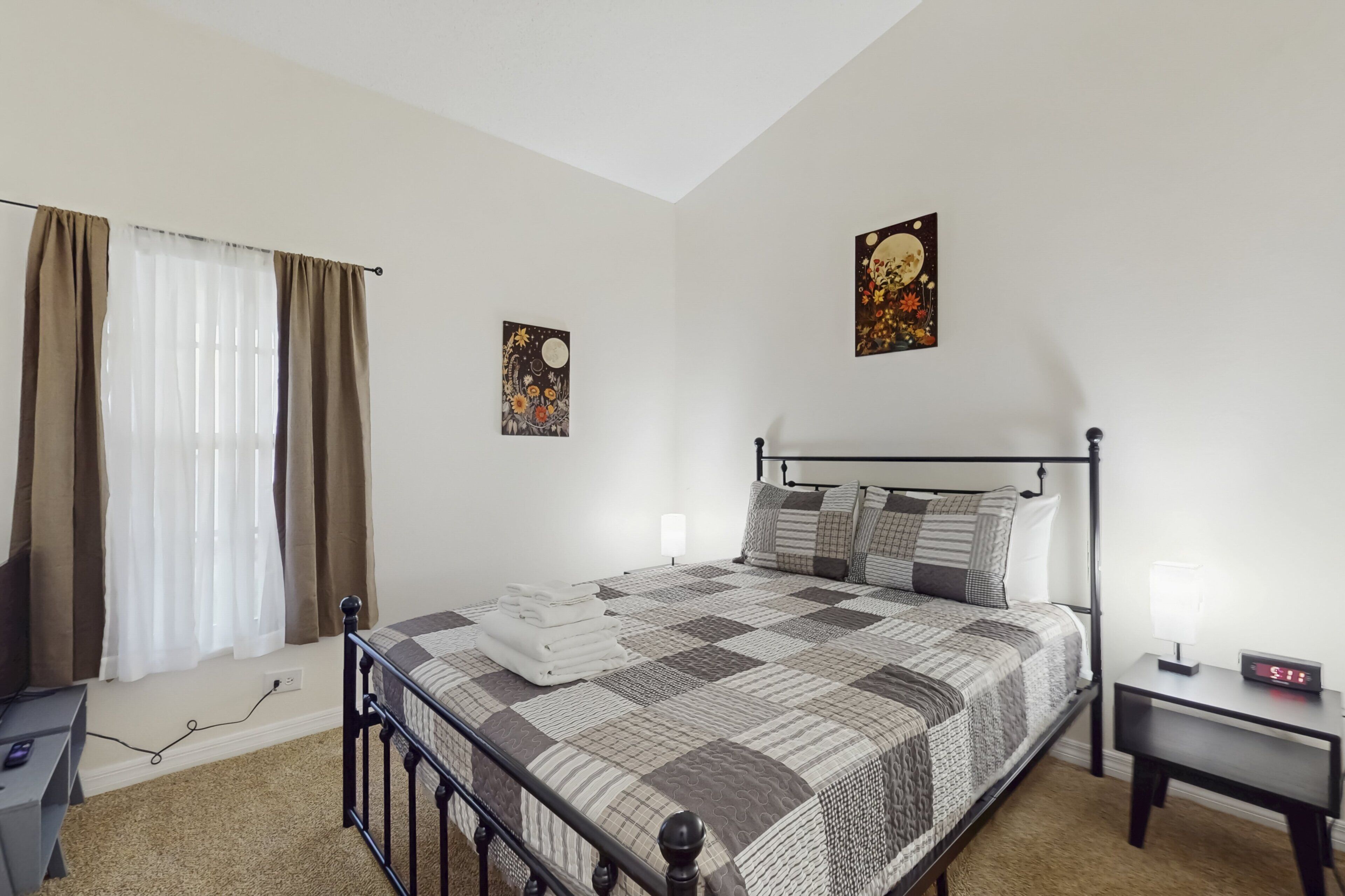A bedroom with a bed, bedside tables, a lamp, a nightstand, a window with curtains, and wall art.