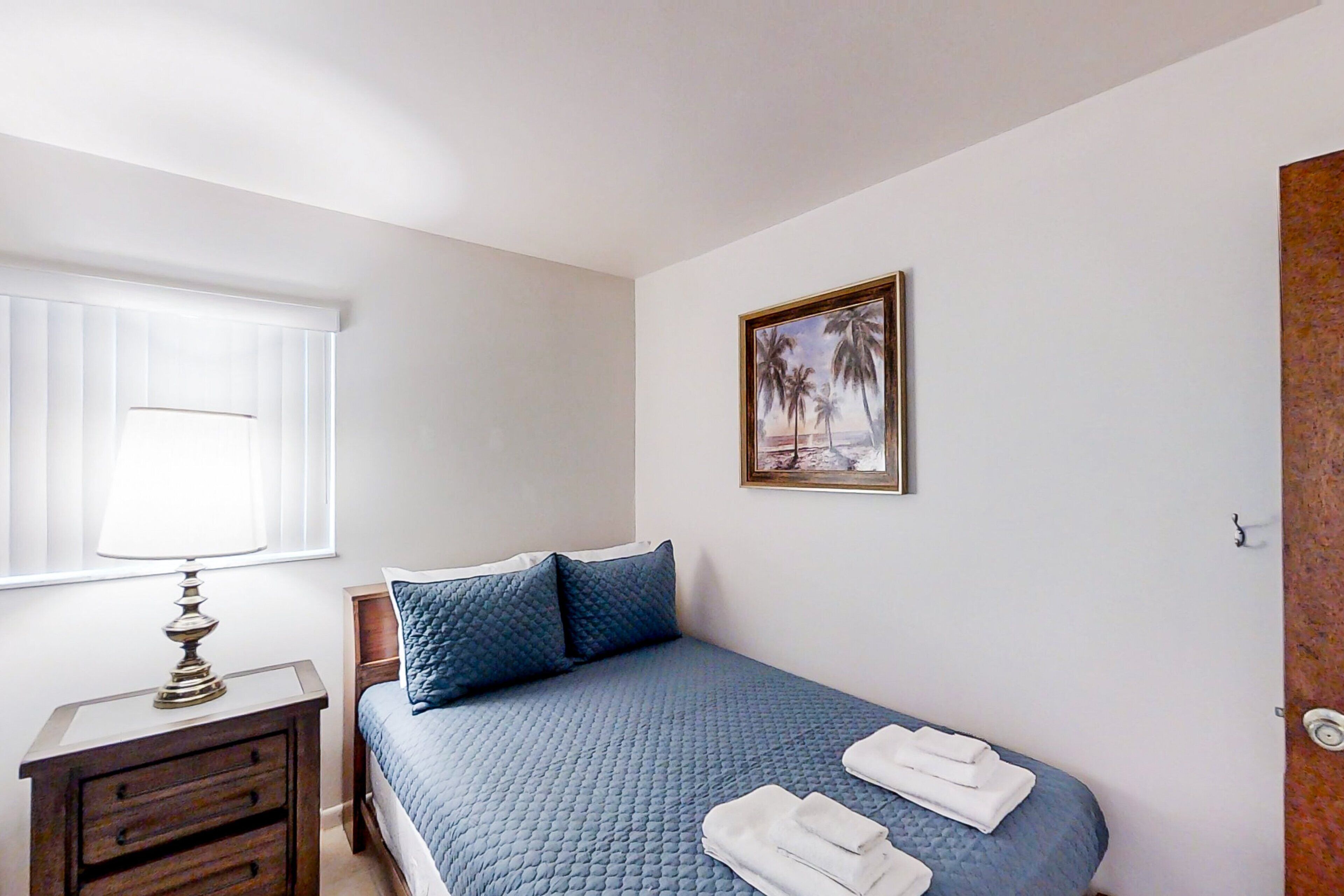 Une chambre à coucher avec un lit, une table de chevet, une lampe, un tableau et une fenêtre avec des stores.
