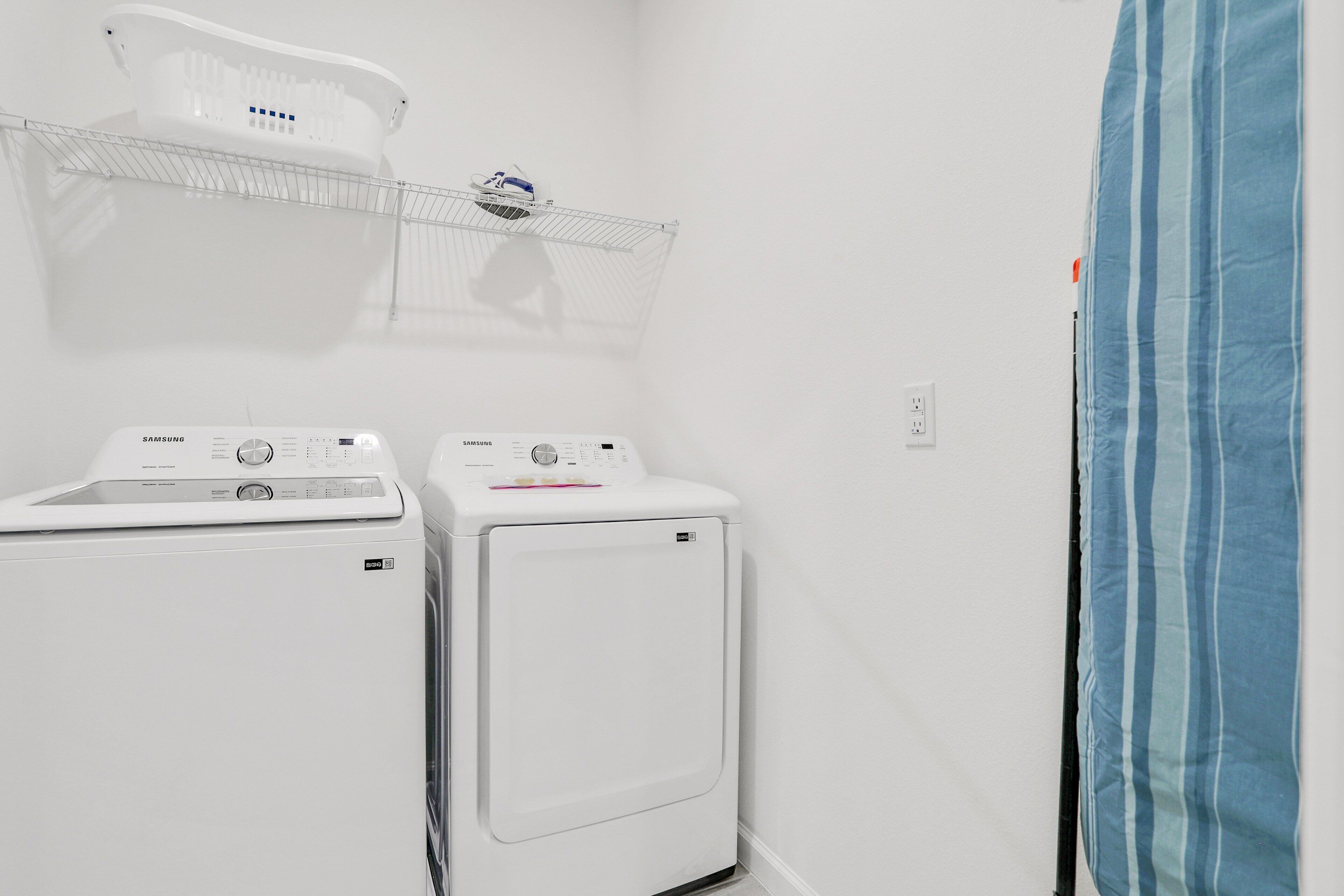 Une buanderie équipée de deux machines à laver et de deux sèche-linge blancs, d’un étendoir métallique et d’un rideau à rayures bleues.