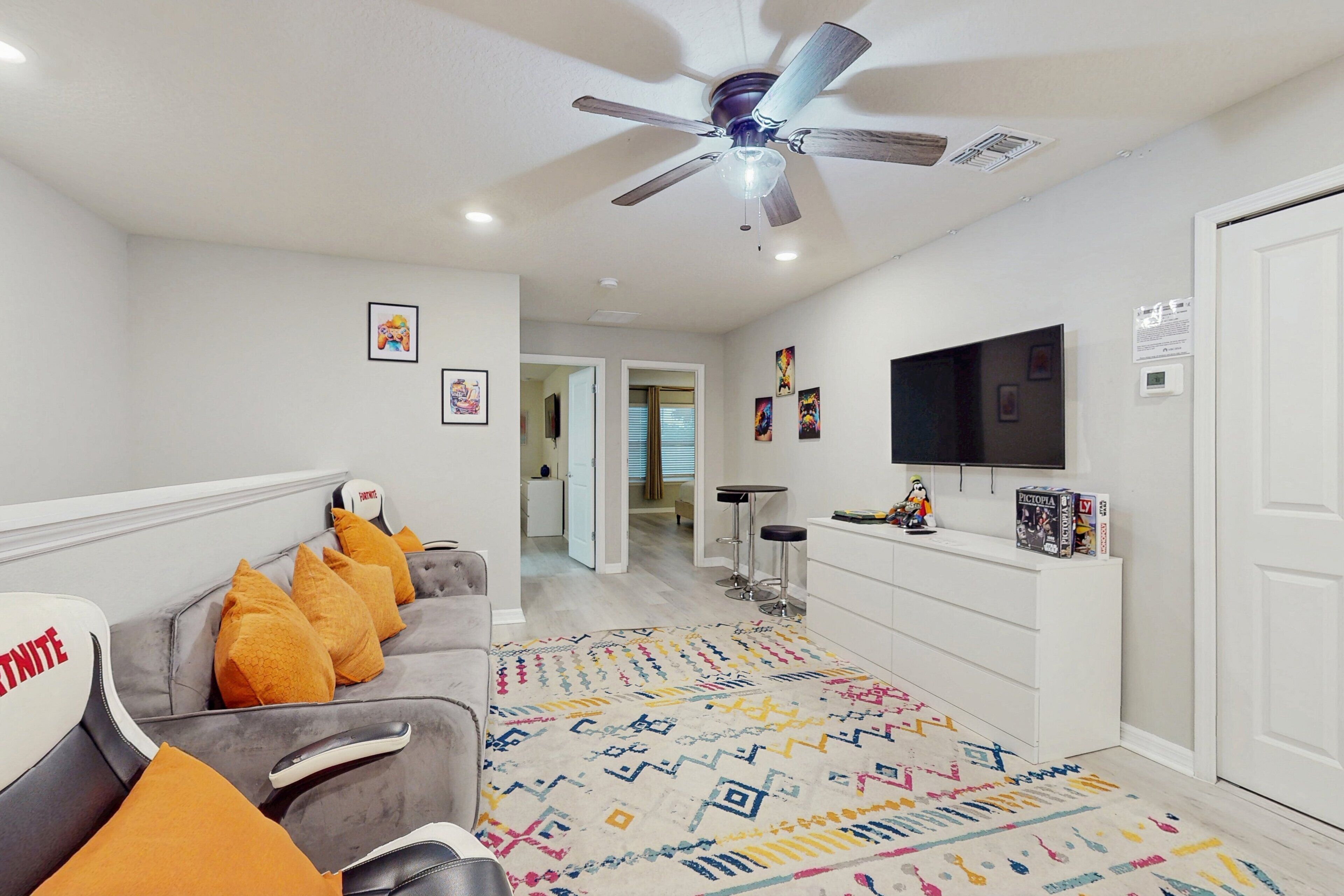 Un salon moderne avec un canapé gris, un tapis coloré, un ventilateur de plafond, une télévision et une porte blanche.