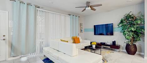 A modern living room with a white sofa, a coffee table, and a TV. There is a ceiling fan, a large potted plant, and a sliding glass door with curtains.