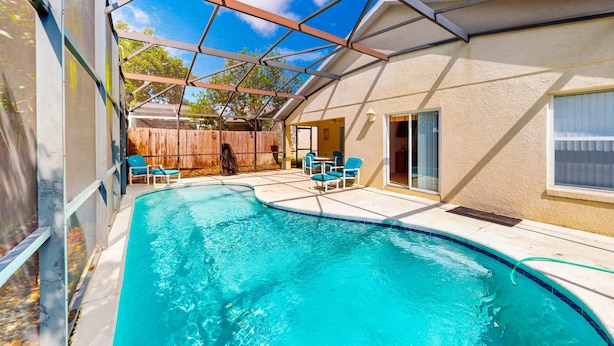 Piscina com um terraço coberto, cadeiras e um cercado de madeira ao fundo.