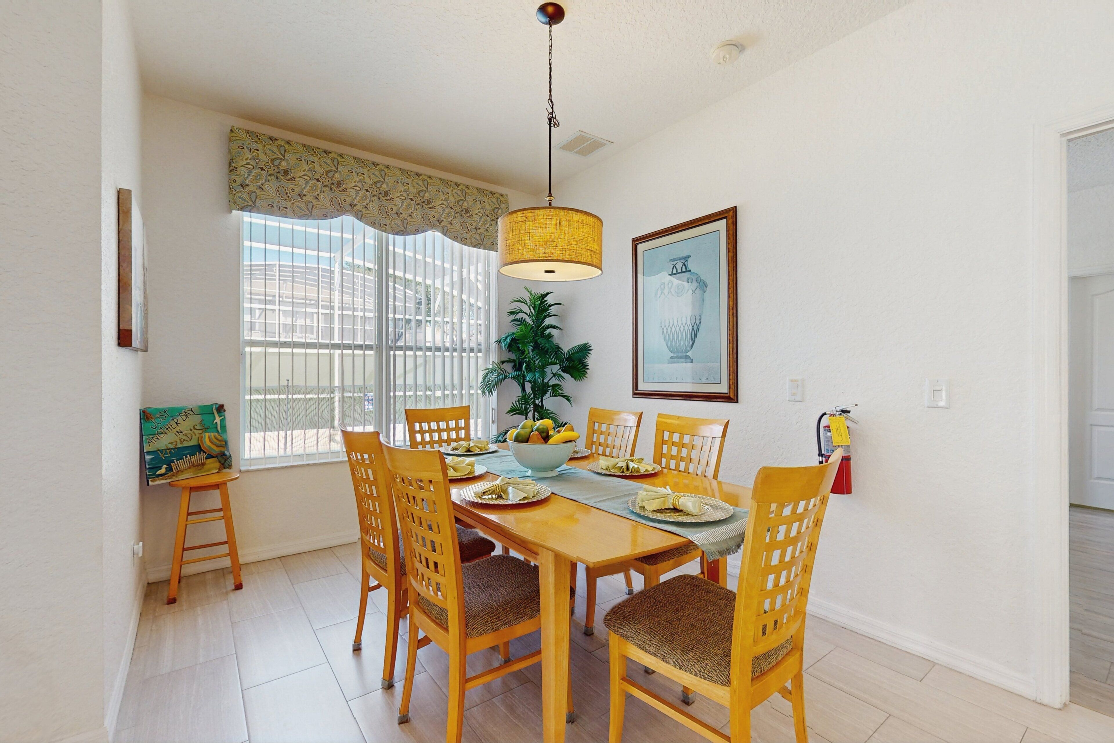 Un coin repas avec une table en bois, des chaises, un luminaire suspendu, une fenêtre avec des stores et un tableau encadré au mur.