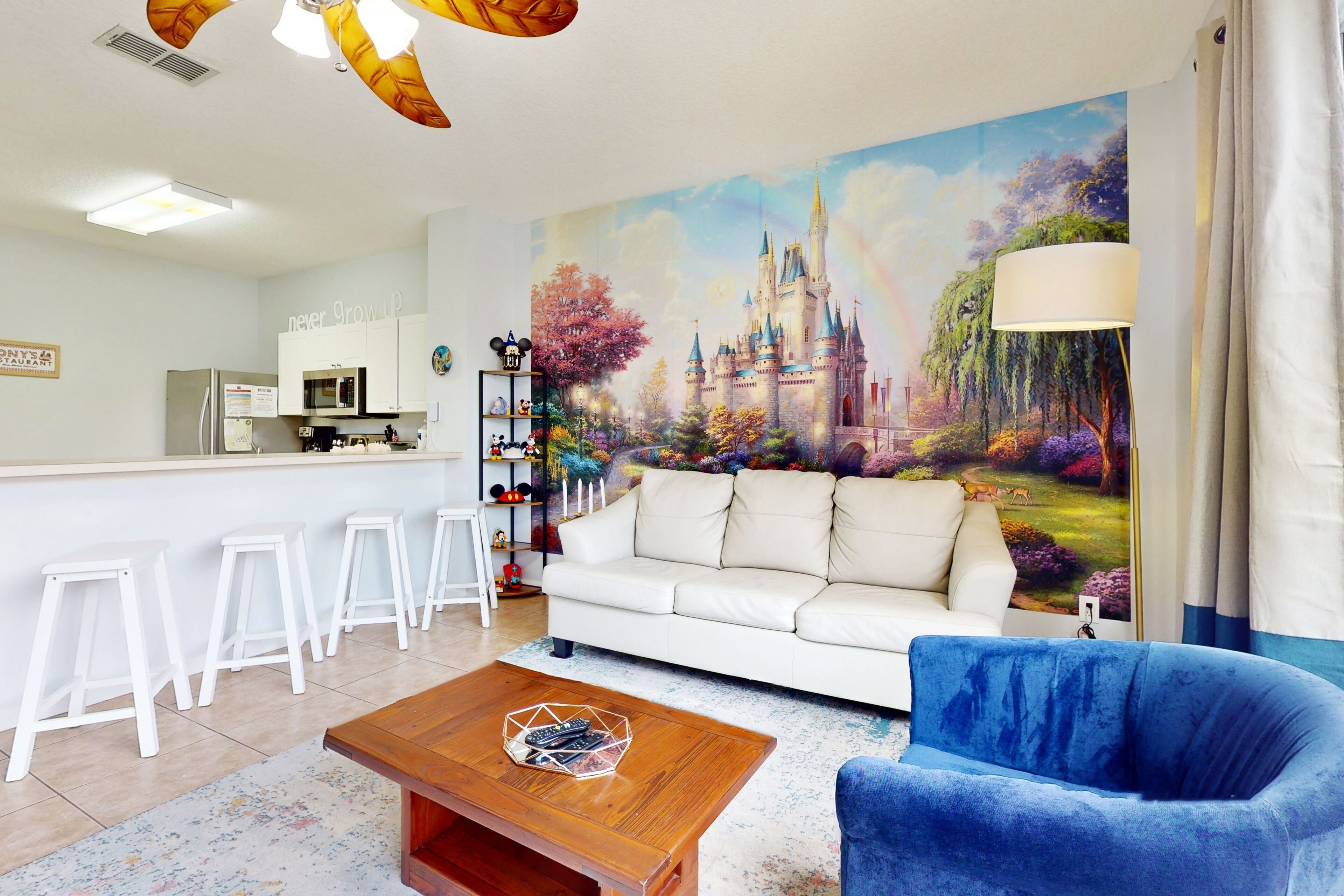 Un salon avec un canapé blanc, un fauteuil bleu et une table basse sur laquelle se trouvent des télécommandes. Un mur est décoré d’une fresque représentant un château, et on y trouve également un coin cuisine avec des meubles blancs et un four à micro-ondes.