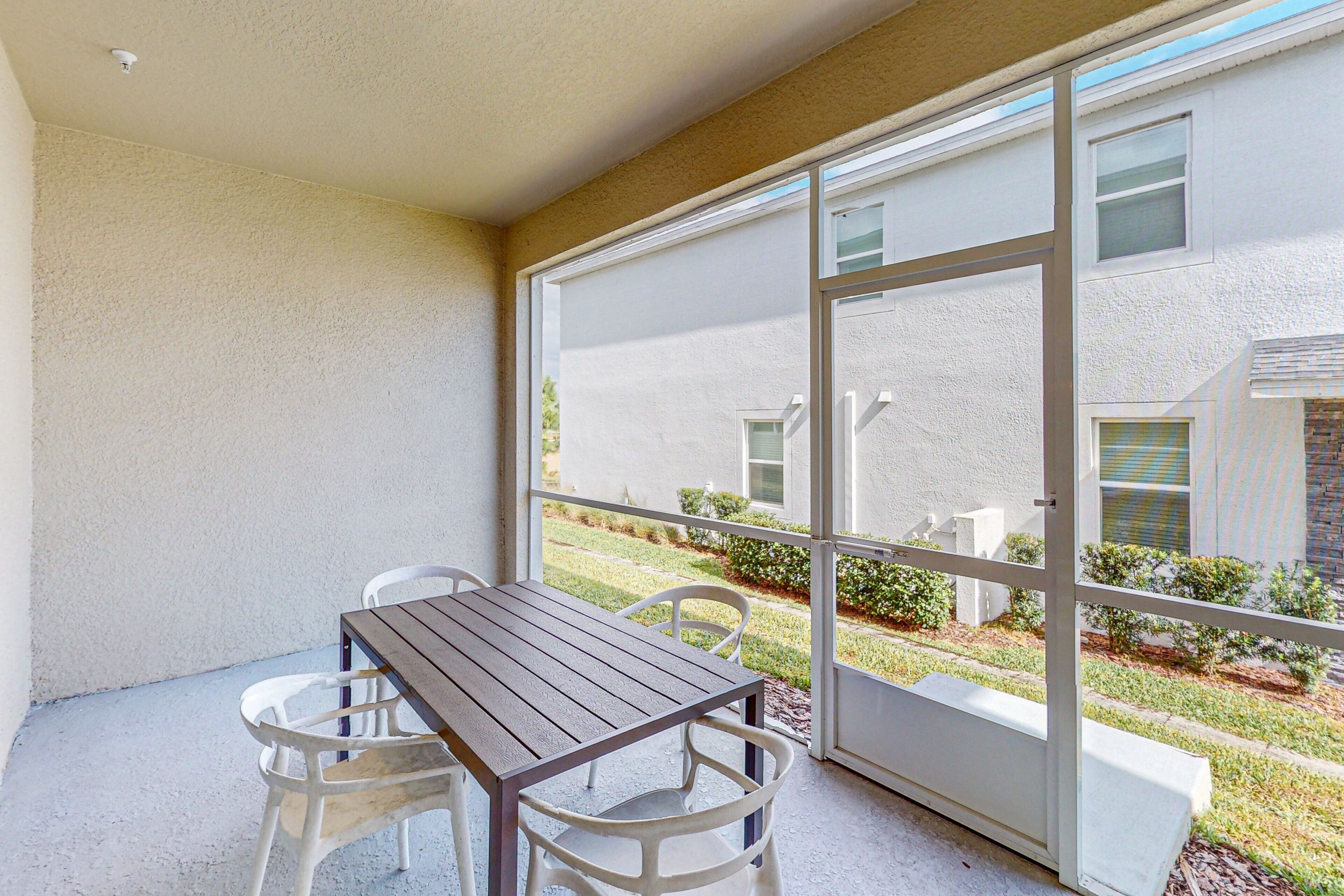 A covered patio with a wooden table and white chairs, large sliding glass doors, and a view of a well-maintained lawn and building.