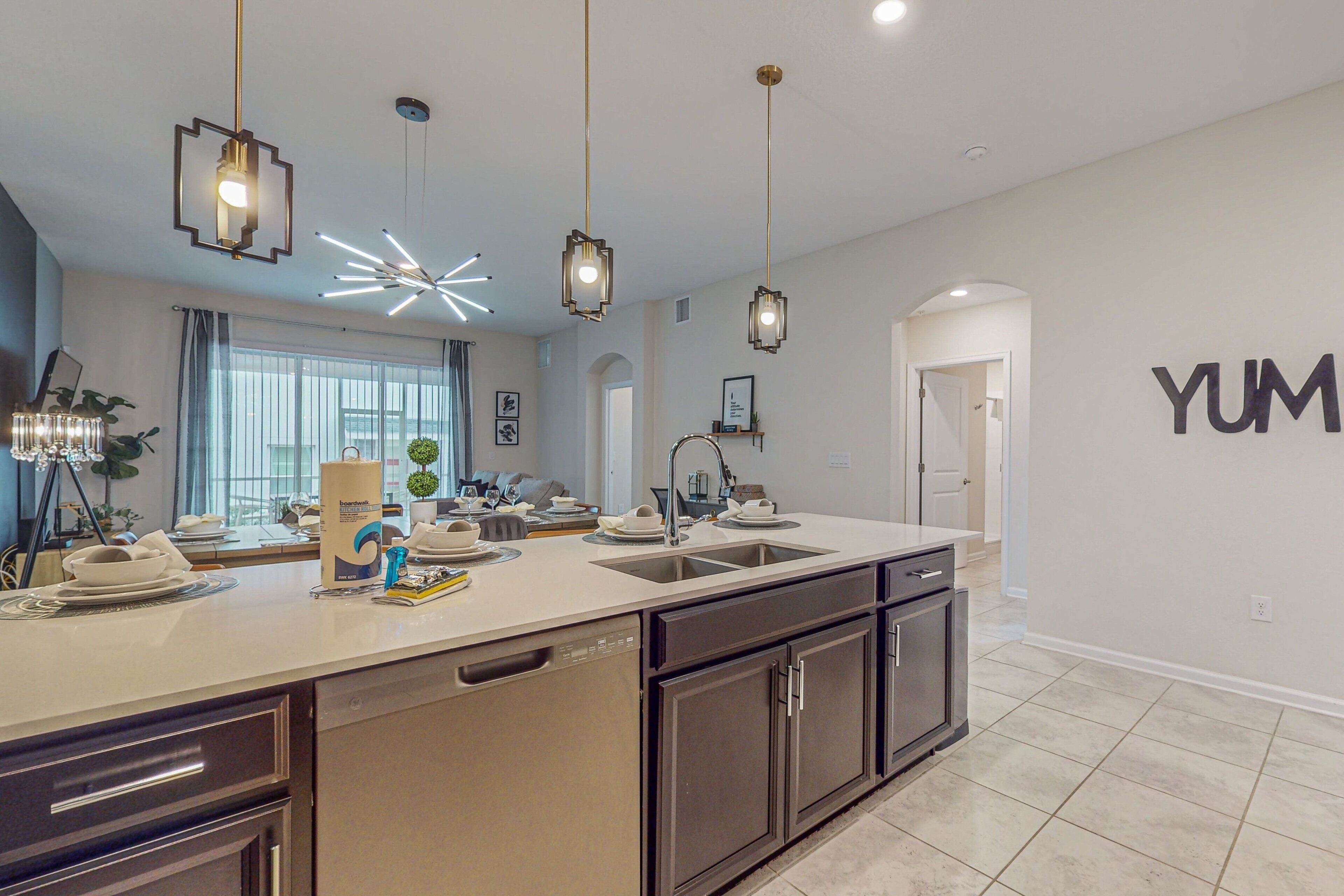 A modern kitchen with a large island, pendant lighting, and a 'YUM' sign on the wall.