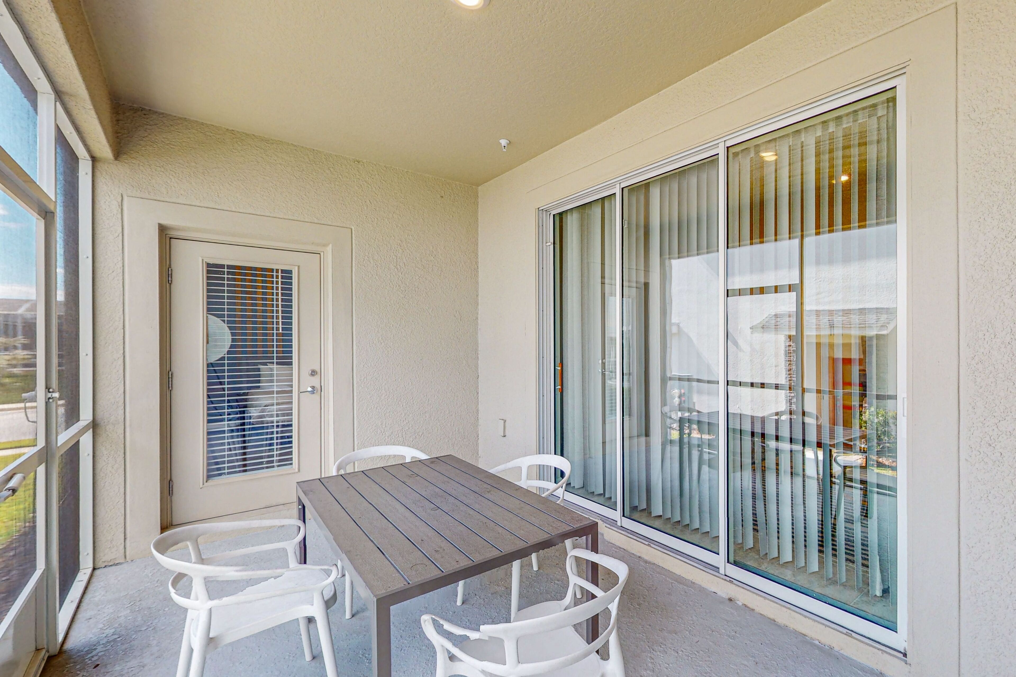 A balcony with a wooden table and white chairs, a sliding glass door, and a view of the outside.