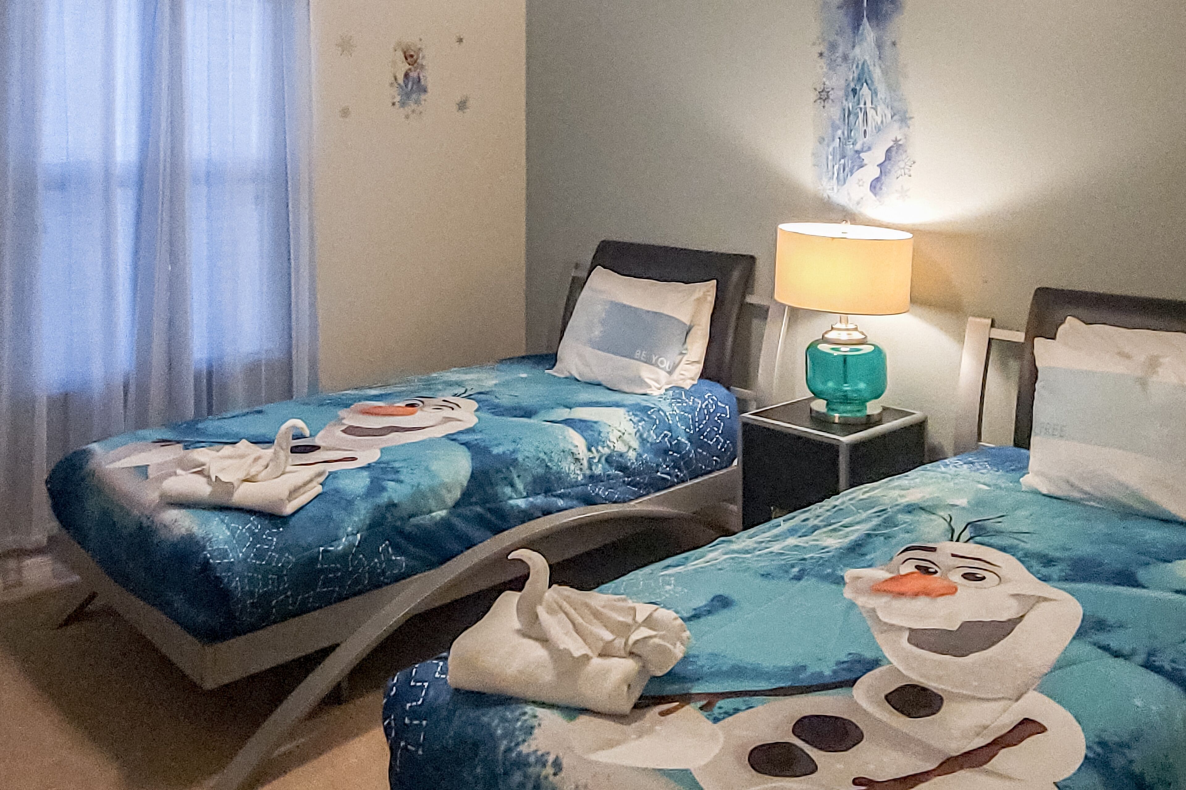 Een hotelkamer met twee bedden, elk voorzien van een blauwe spreidek met een sneeuwpop ontwerp. Tussen de bedden staat een nachtkastje met een lamp.