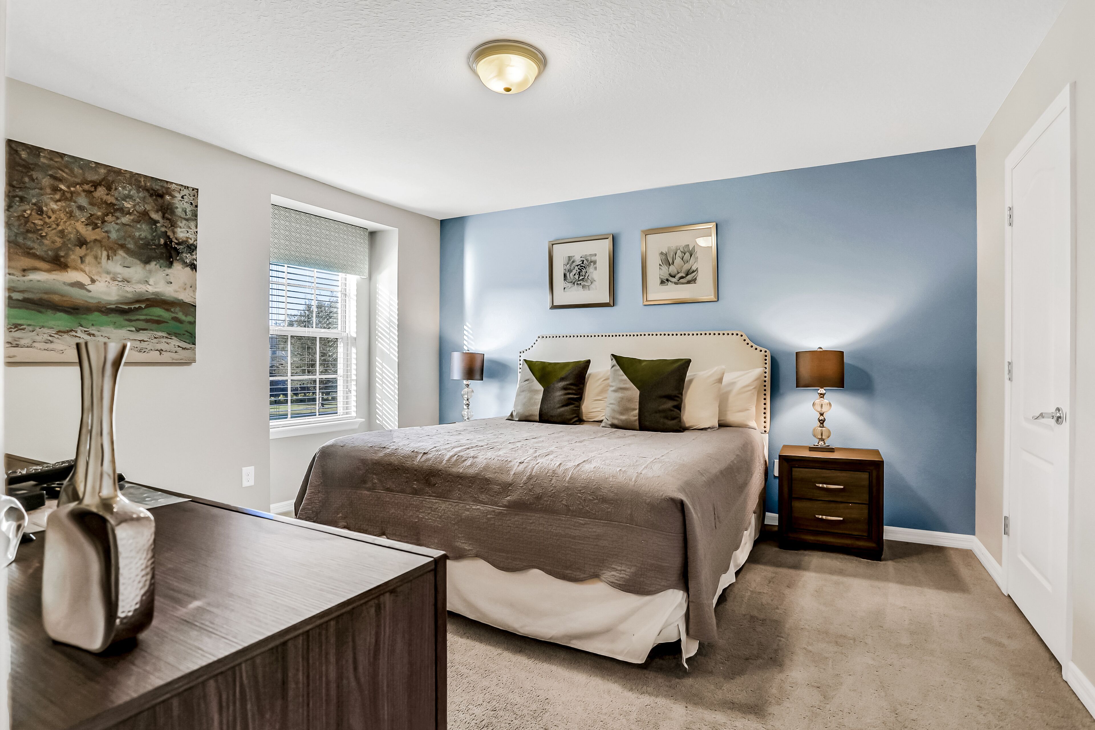 A bedroom with a large bed, a desk, a lamp, and artwork on the wall.